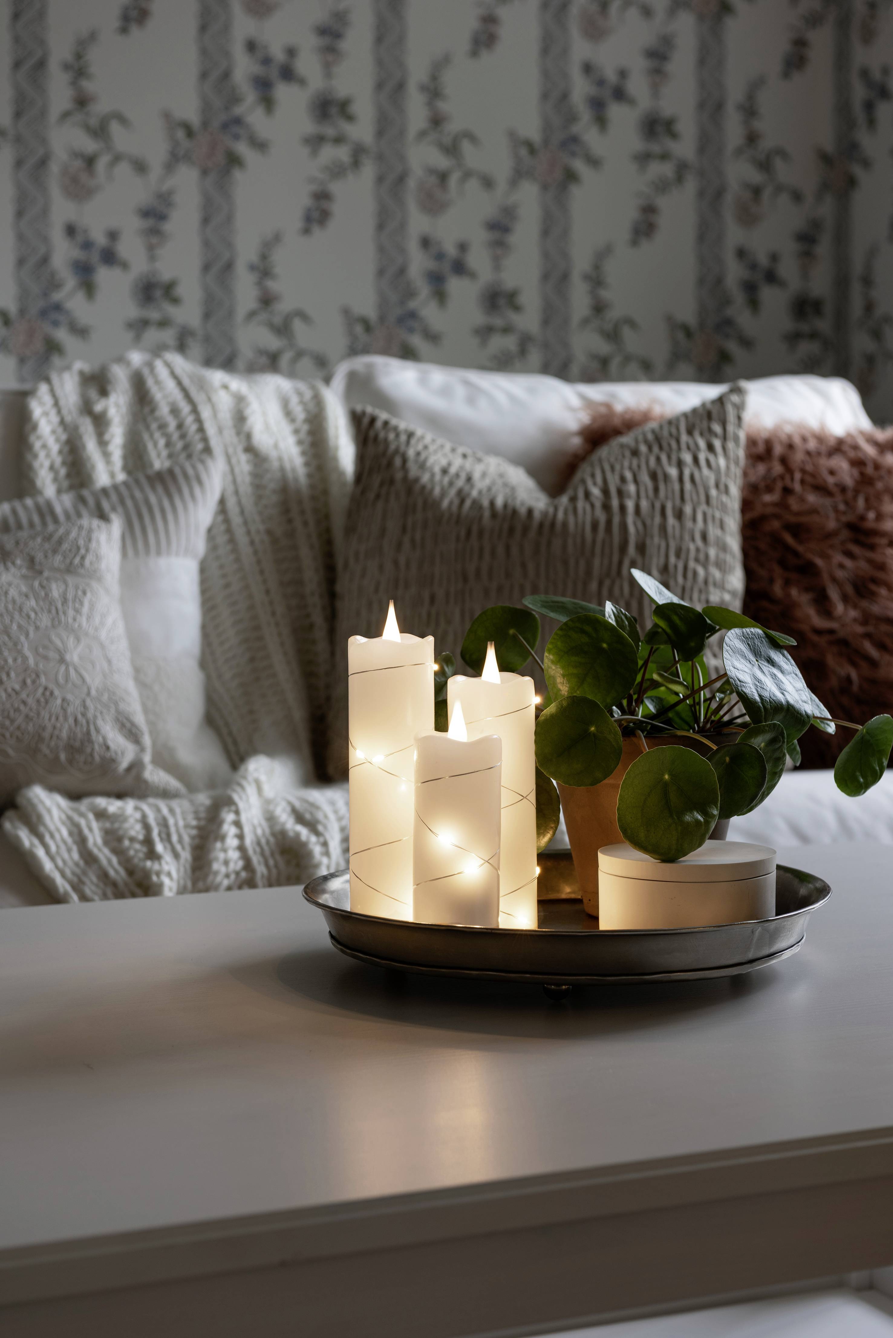 Przytulny salon z białymi świecami i rośliną na stole; w tle kanapa z poduszkami na tle wzorzystej tapety.