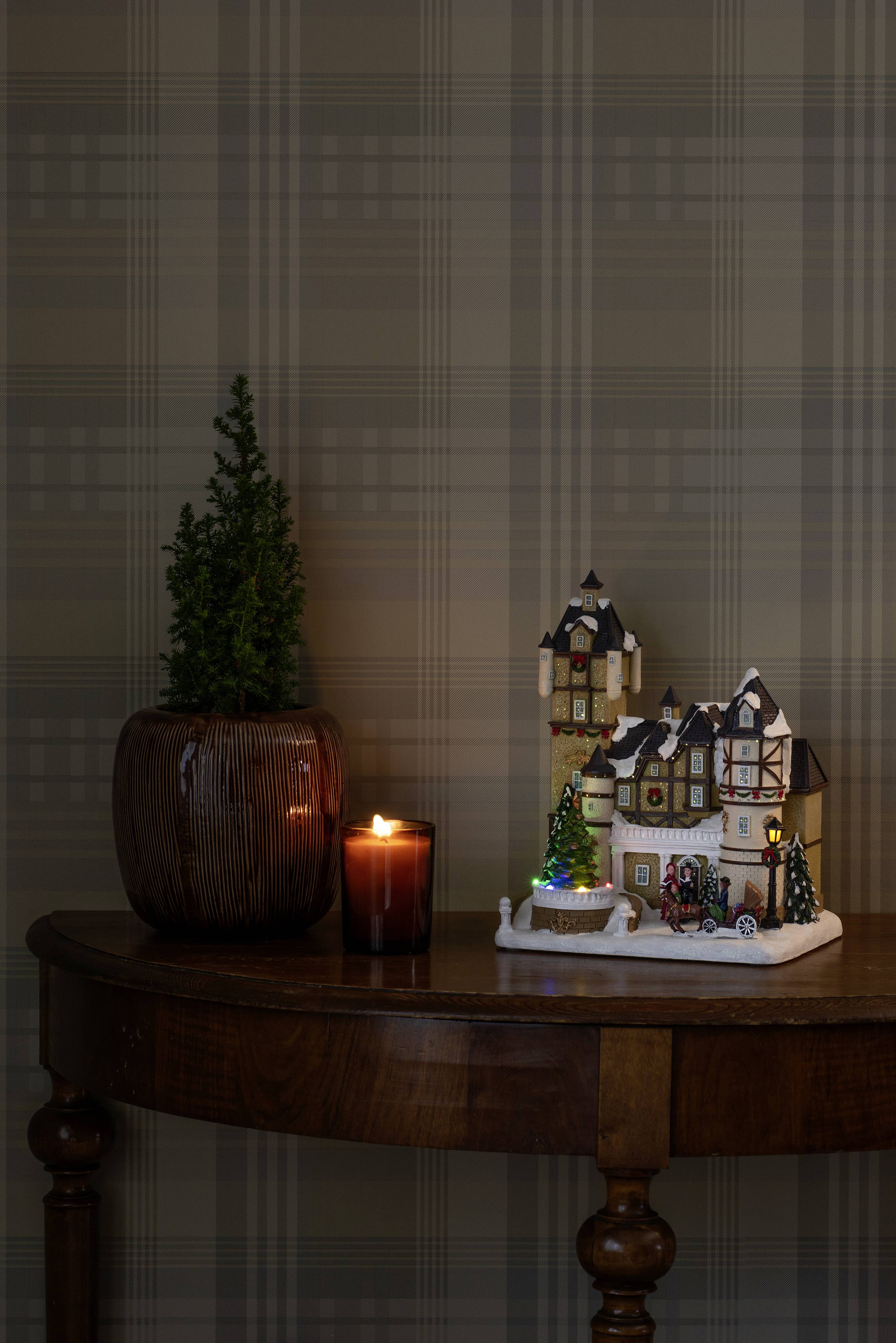 Świecznik z miniaturową wioską bożonarodzeniową i rośliną, ciepło oświetlony, na drewnianym stole na tle we wzory.