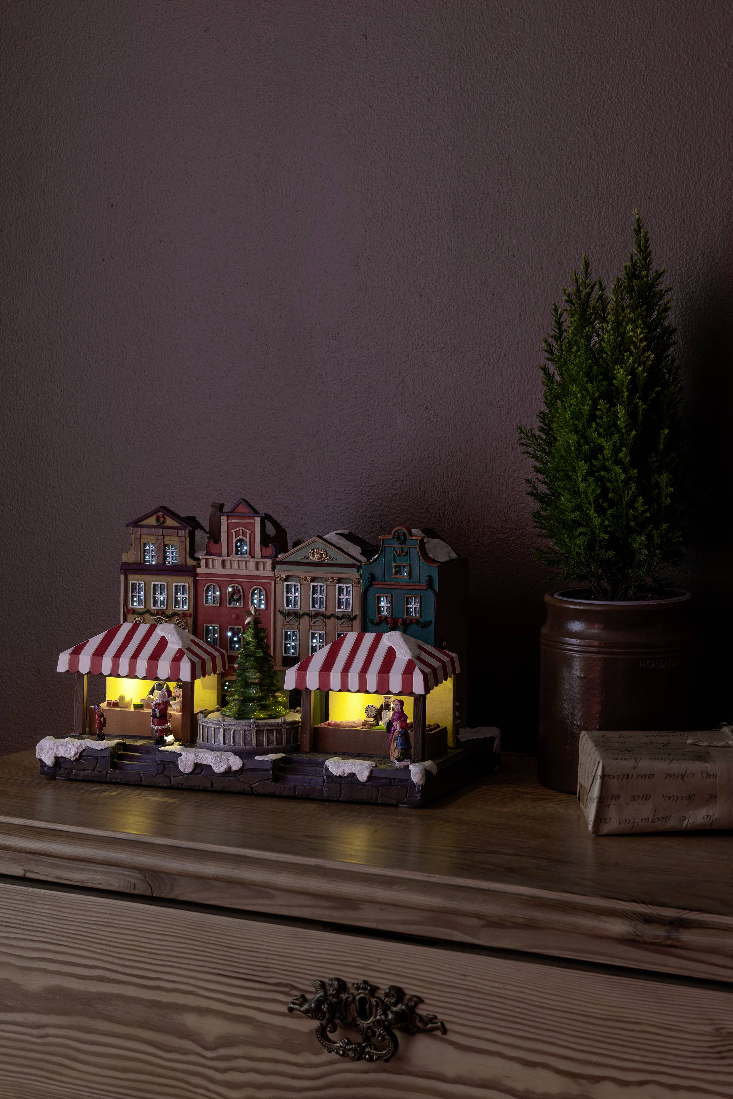 Oświetlona miniaturowa wioska bożonarodzeniowa z kolorowymi domkami i małymi figurkami stoi na komodzie. Obok znajduje się mała choinka doniczkowa.