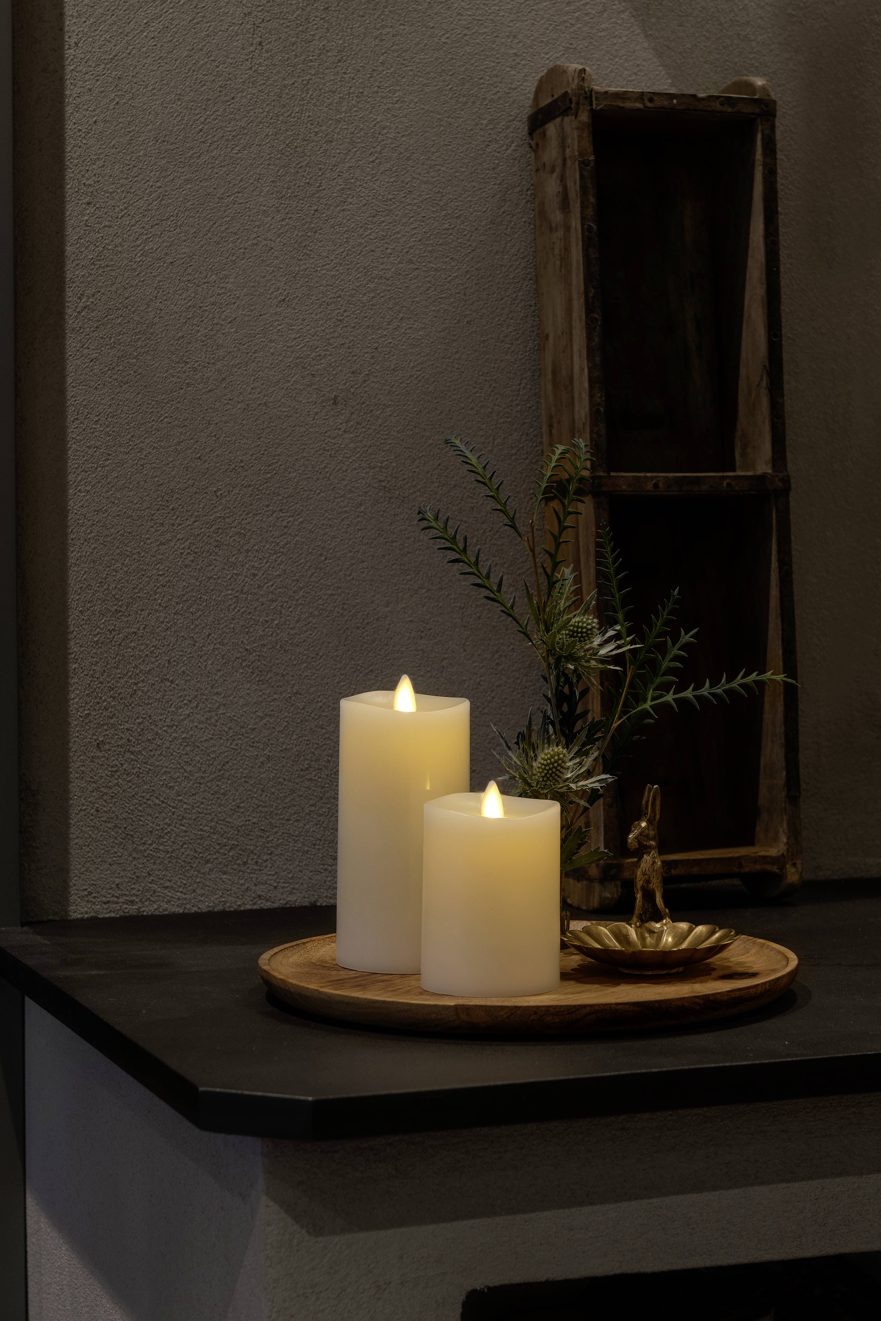 Dwie płonące, białe świece na okrągłej drewnianej tacy, obok małej gałązki i złotego elementu dekoracyjnego na półce.
