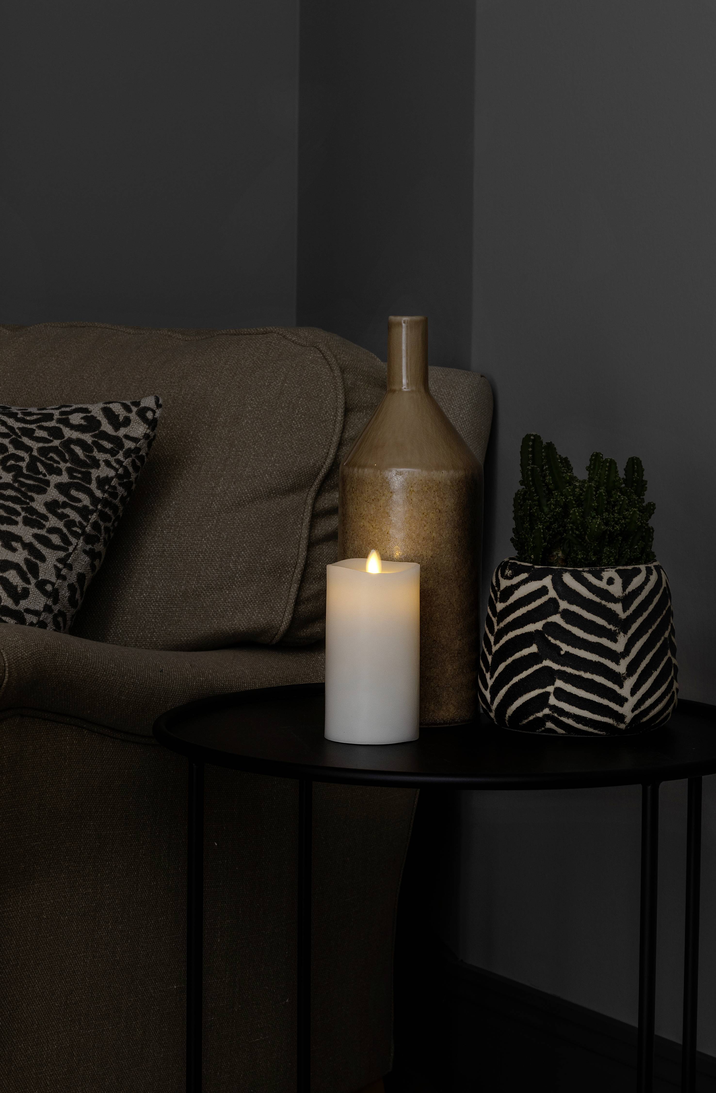 Ciemny salon z brązową kanapą, ozdobną poduszką, białymi świecami na stole i doniczkową rośliną w doniczce w zebrę.