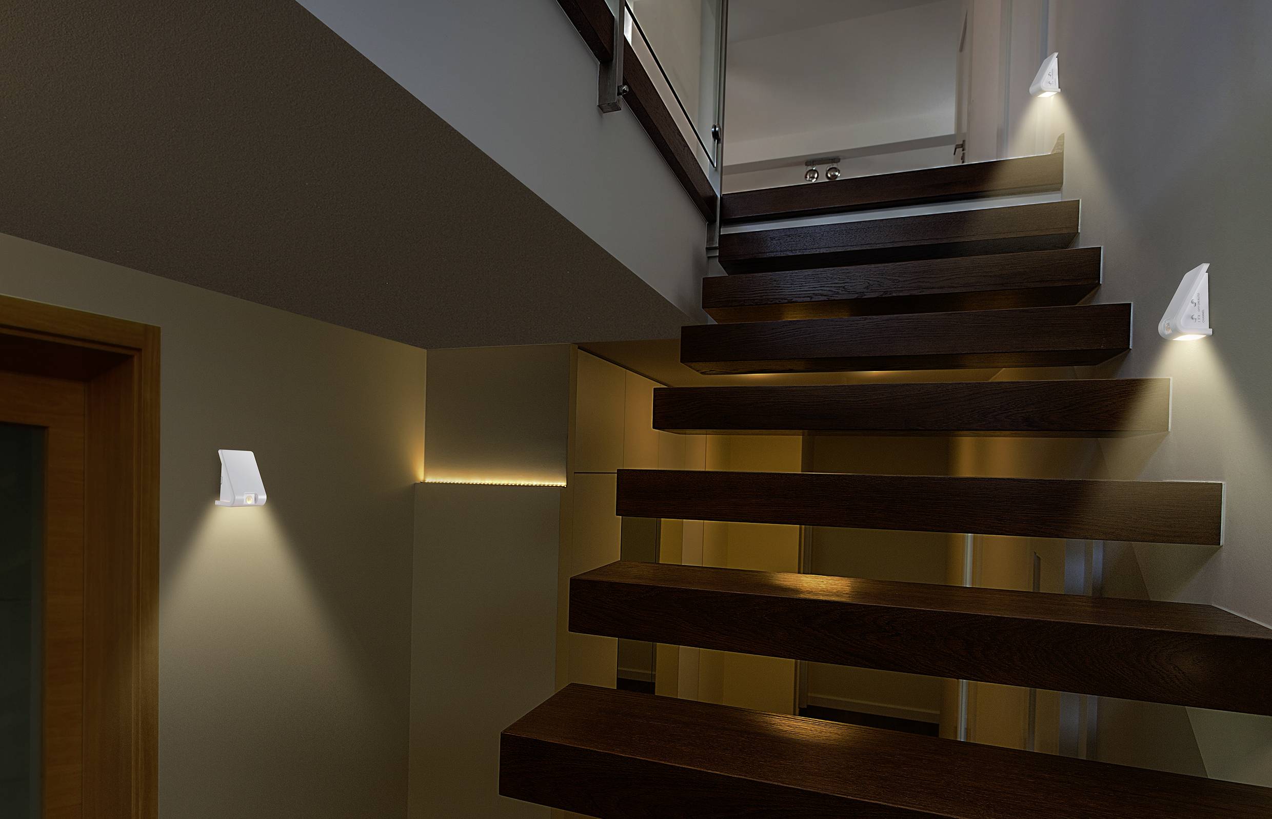 Drewniane schody w ciemnym kolorze we wnętrzu w stylu nowoczesnym, oświetlone kinkietami po obu stronach, rzucającymi ciepłe światło na stopnie.