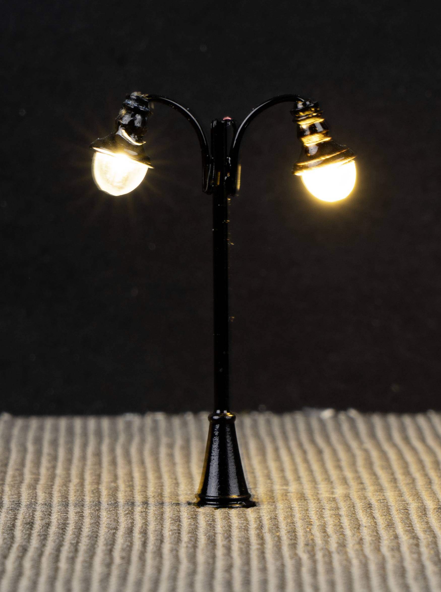 Mała, czarna latarnia uliczna z dwoma świecącymi żarówkami stoi na strukturalnym podłożu na ciemnym tle.