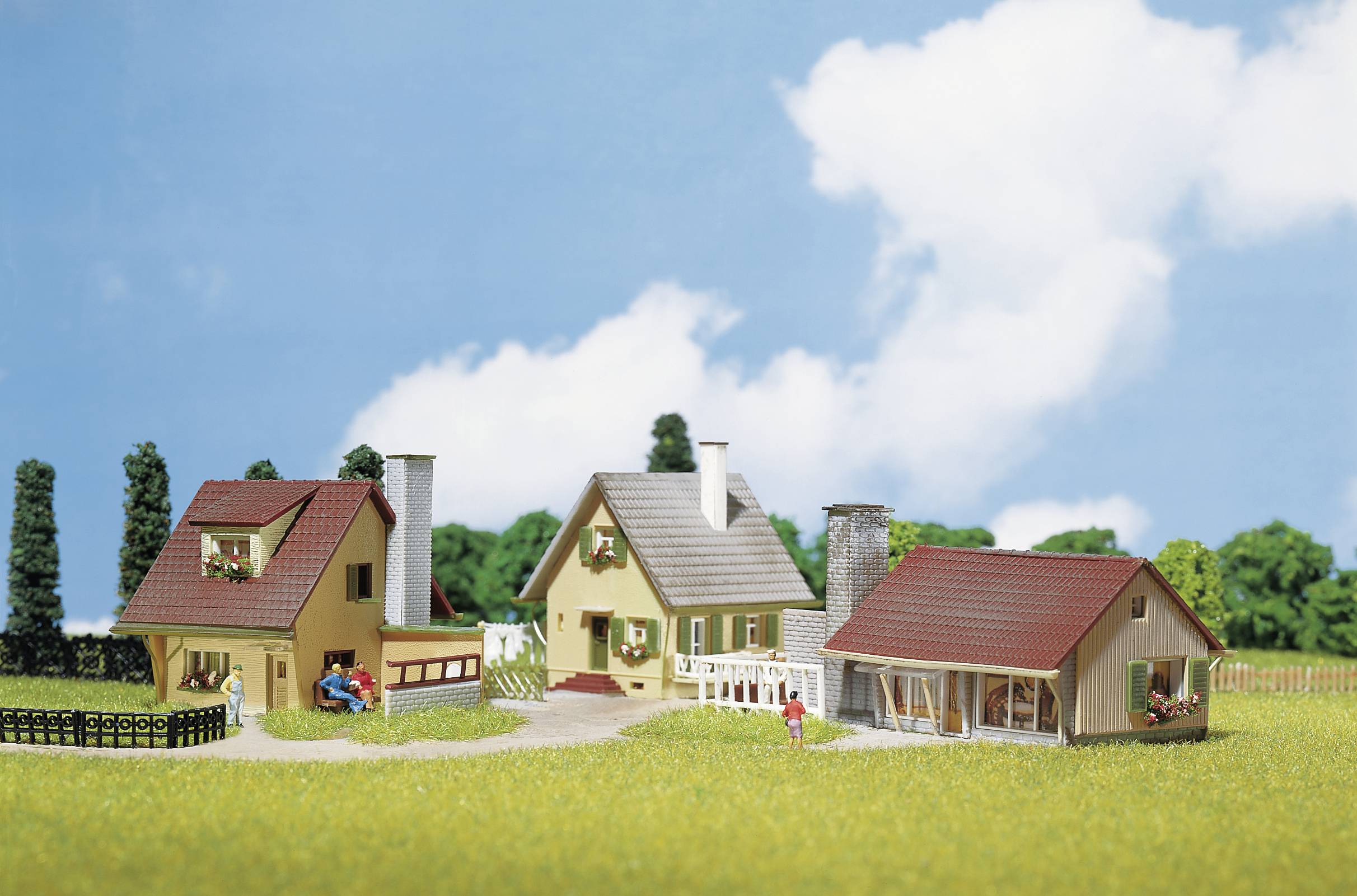 Trzy modelowe domy w wiejskim otoczeniu, otoczone drzewami i łąką, pod błękitnym niebem z kilkoma białymi chmurami.