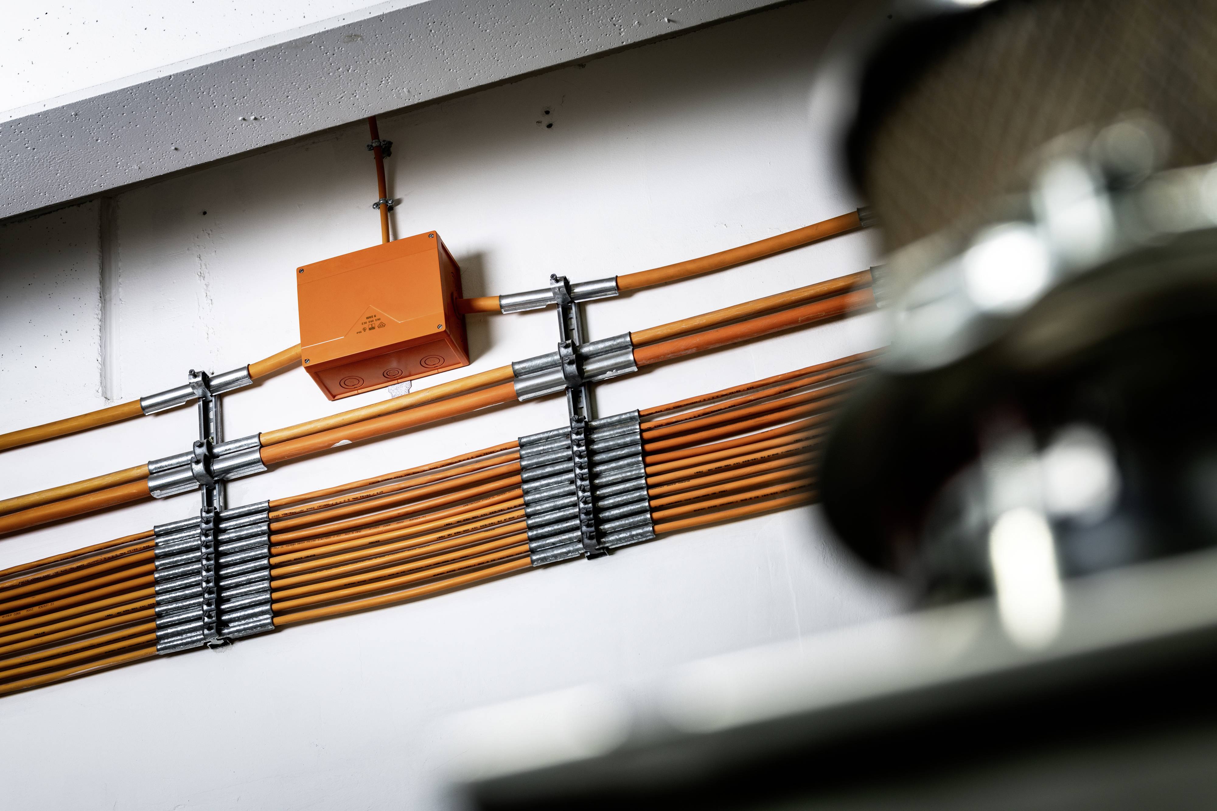 Pomarańczowe kable biegną starannie wzdłuż ściany. Pomarańczowe pudełko jest przymocowane do kabli. Metalowe komponenty są nieostre na pierwszym planie.