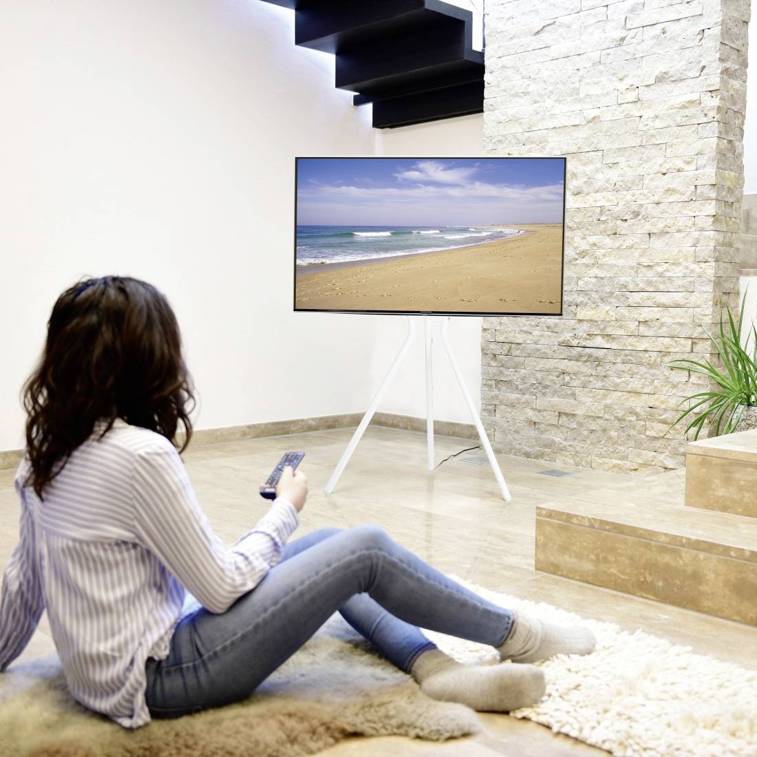 Kobieta siedzi na dywanie w salonie i ogląda telewizję. Na ekranie widać krajobraz plaży. Trzyma w ręku pilot.