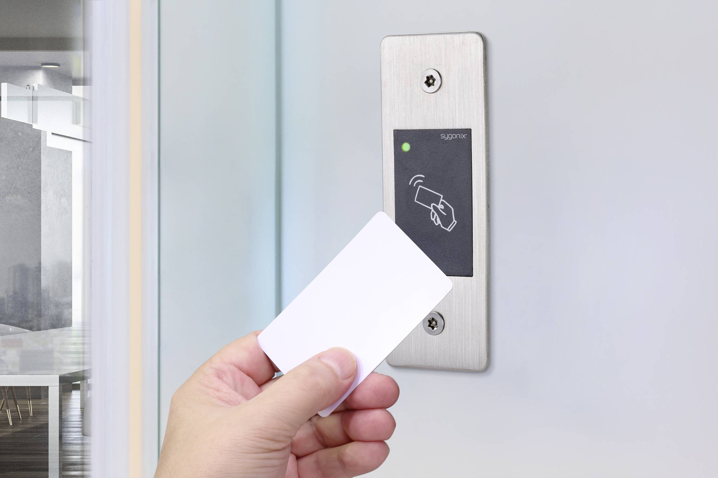 Ręka trzyma kartę dostępową przed czytnikiem przy ścianie, emitującym zielone światło, w nowoczesnym budynku biurowym.