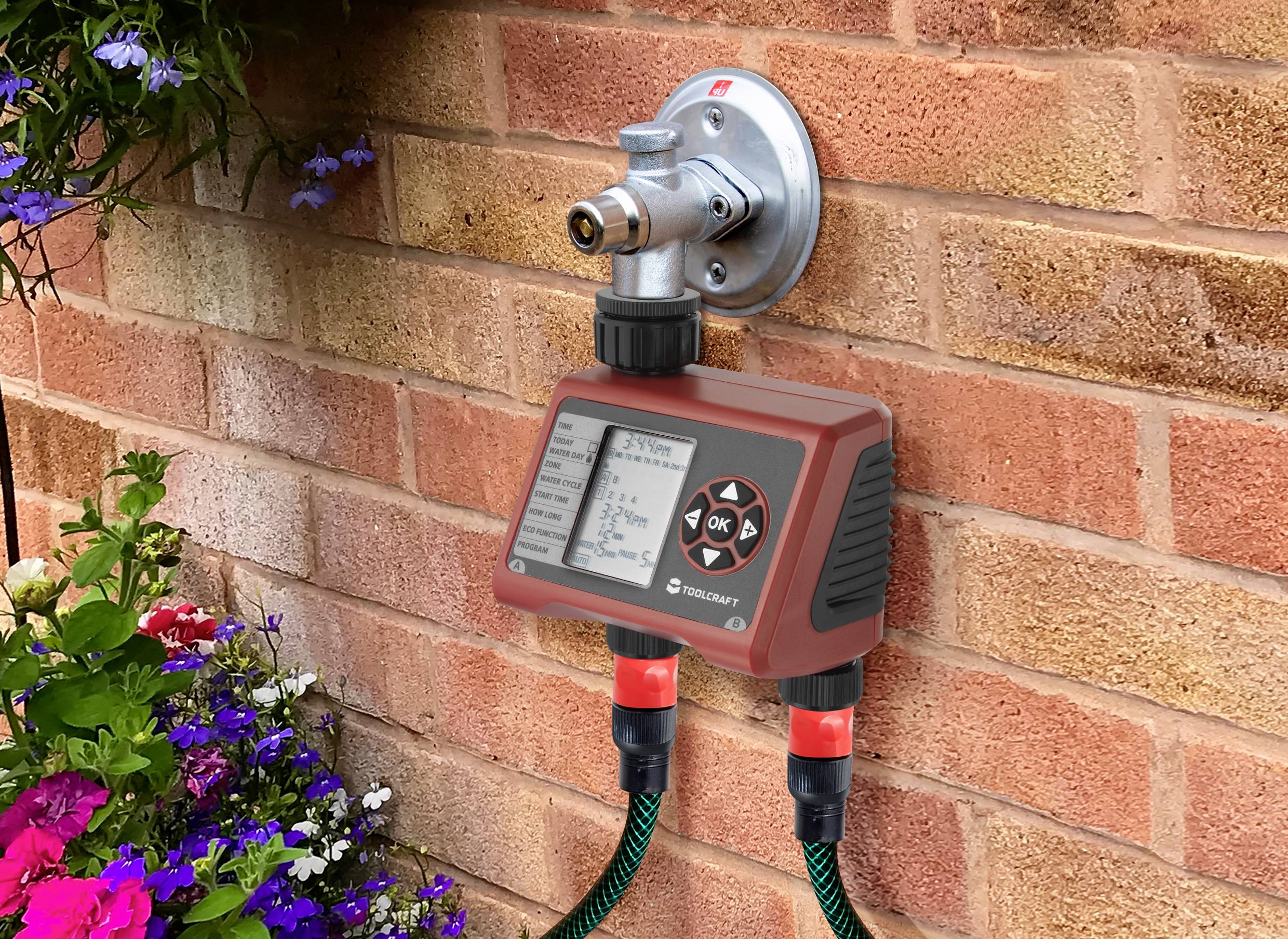 Kran z cyfrowym urządzeniem sterującym nawadnianiem na ceglastej ścianie. Poniżej znajdują się dwa węże ogrodowe i rabata kwiatowa.