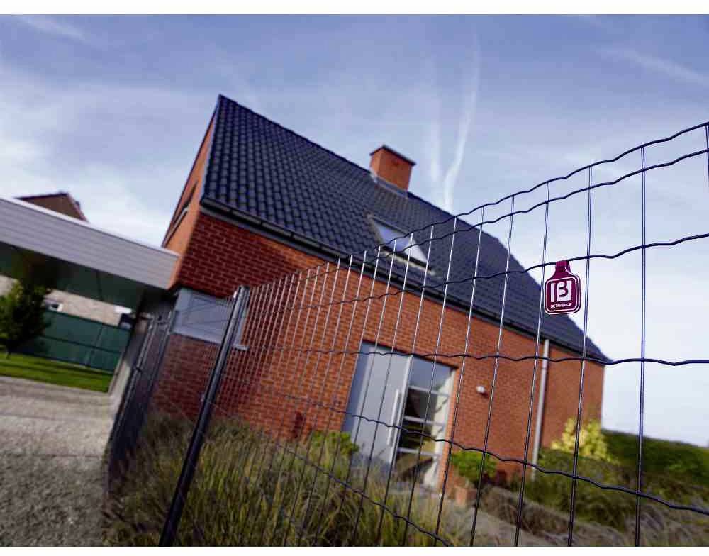 Czerwony dom z cegły z ukośnym dachem za płotem z siatki drucianej. Niebo jest błękitne, a po lewej stronie znajduje się wiata samochodowa.