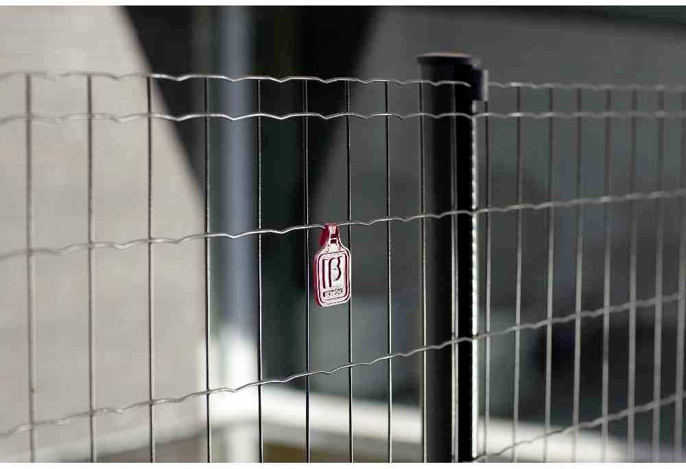 Czerwona plomba zabezpieczająca jest przymocowana do ogrodzenia drucianego. W tle widać nieostre budynki.