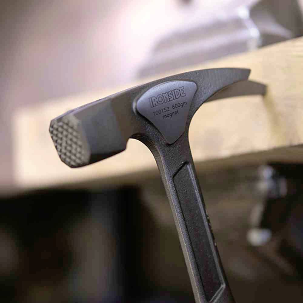 Młotek leży na drewnianym bloku. Jest podtrzymywany przez metalowy pręt. Ostrość obrazu skupiona jest na prążkowanej powierzchni młotka.