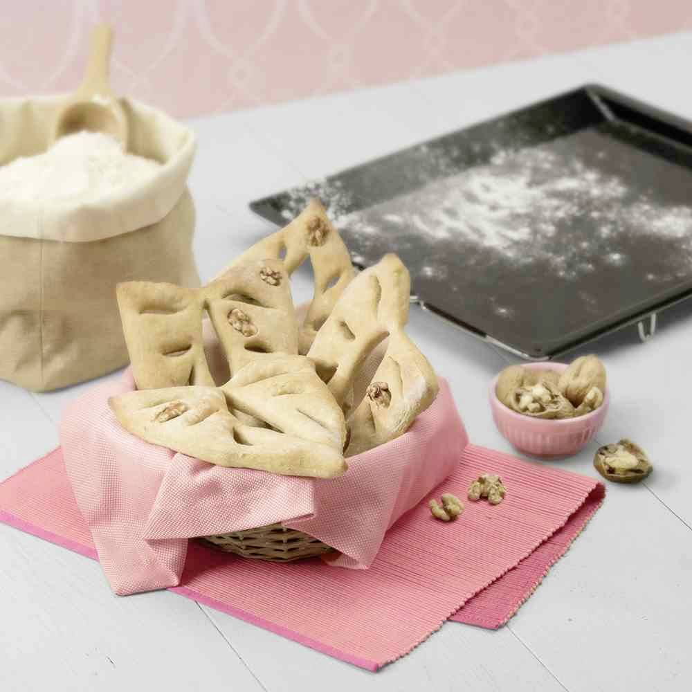 Kosz z wypieczonymi drożdżówkami, ozdobionymi orzechami włoskimi, stoi na różowej serwecie. Obok leżą mąka i pusta blacha do pieczenia.