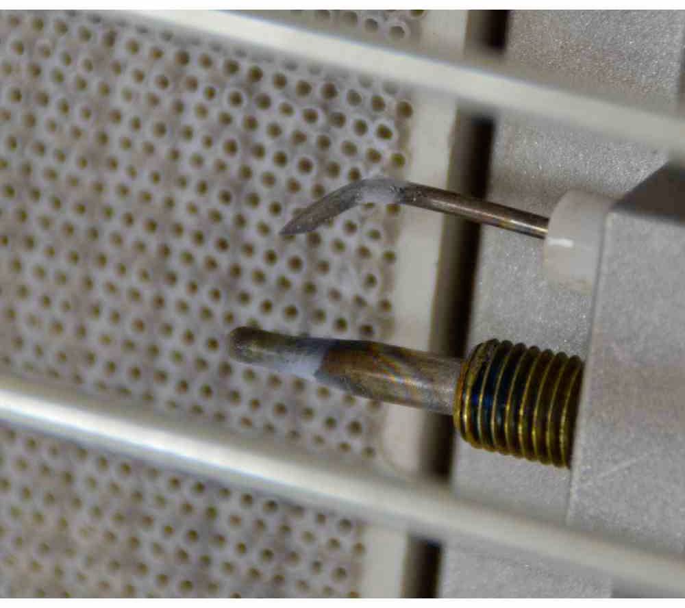 Zbliżenie uszkodzonego grzejnika z pękniętą spiralą grzewczą wystającą z ceramicznej obudowy. Tło przedstawia drobnooka kratka.