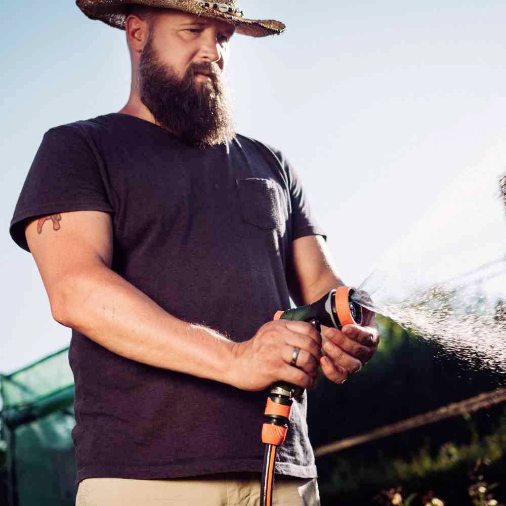 Mężczyzna w słomkowym kapeluszu podlewa rośliny wężem ogrodowym w słoneczny dzień.