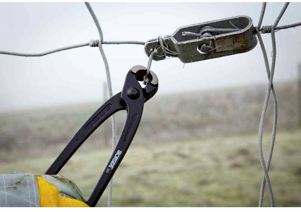 Jedna osoba przecina siatkę ogrodzeniową za pomocą szczypiec do cięcia prętów. Otoczenie jest mgliste i przypomina wiejski krajobraz.