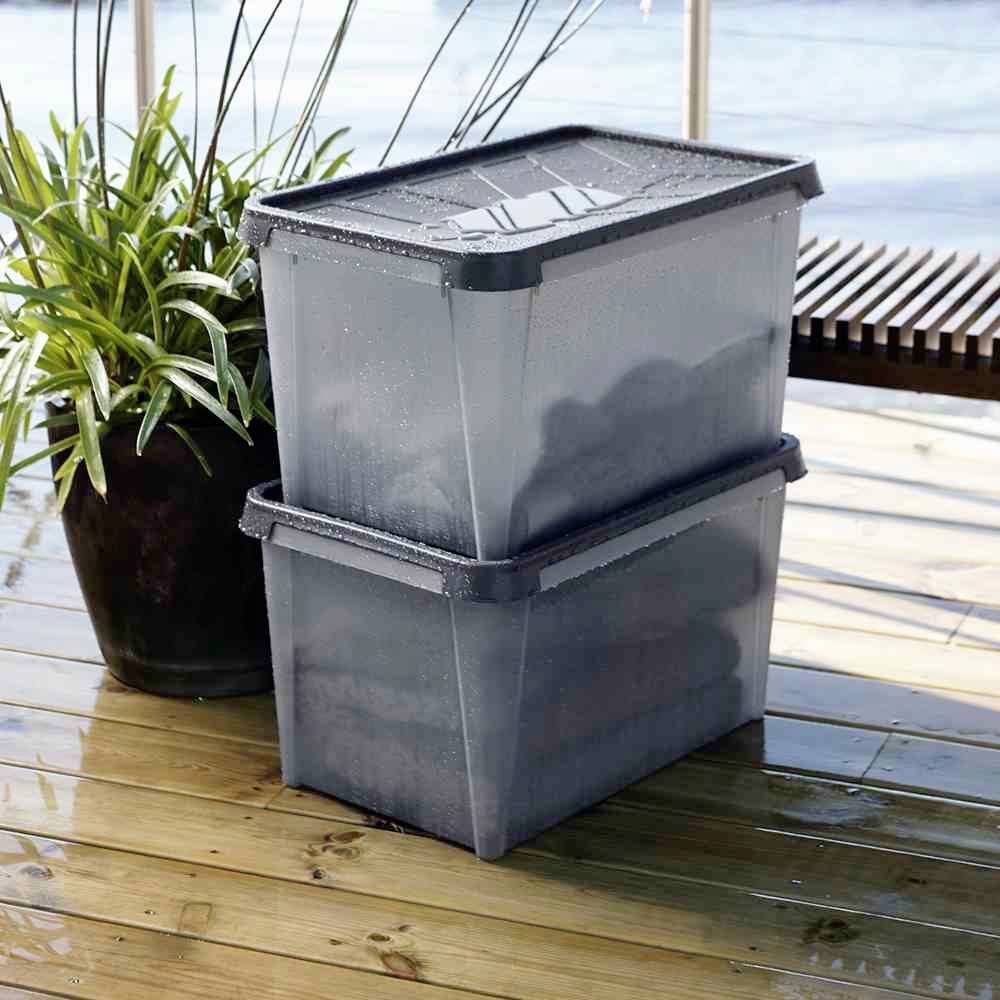 Dwie ułożone na sobie szare plastikowe skrzynki na wilgotnej drewnianej podłodze obok rośliny, z kroplami wody na ich powierzchni.