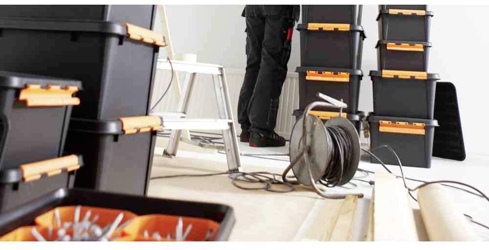 Robotnik na drabinie układa czarne skrzynki narzędziowe z pomarańczowymi zamknięciami w pomieszczeniu wypełnionym materiałami budowlanymi.