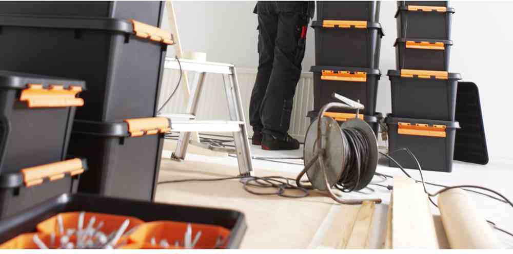 Osoba pracuje w pomieszczeniu, otoczona ułożonymi stosami czarnych skrzynek narzędziowych z pomarańczowymi zamknięciami. Na podłodze leży szpula kablowa.