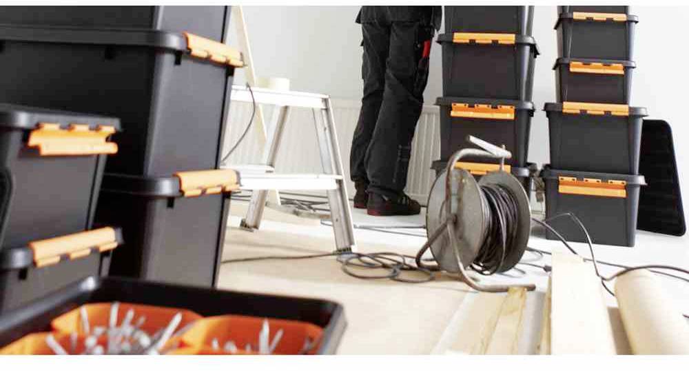 Robotnik stoi pomiędzy ułożonymi w stos czarnymi skrzynkami narzędziowymi z pomarańczowymi zamknięciami w pomieszczeniu z narzędziami budowlanymi i rolką kabli.