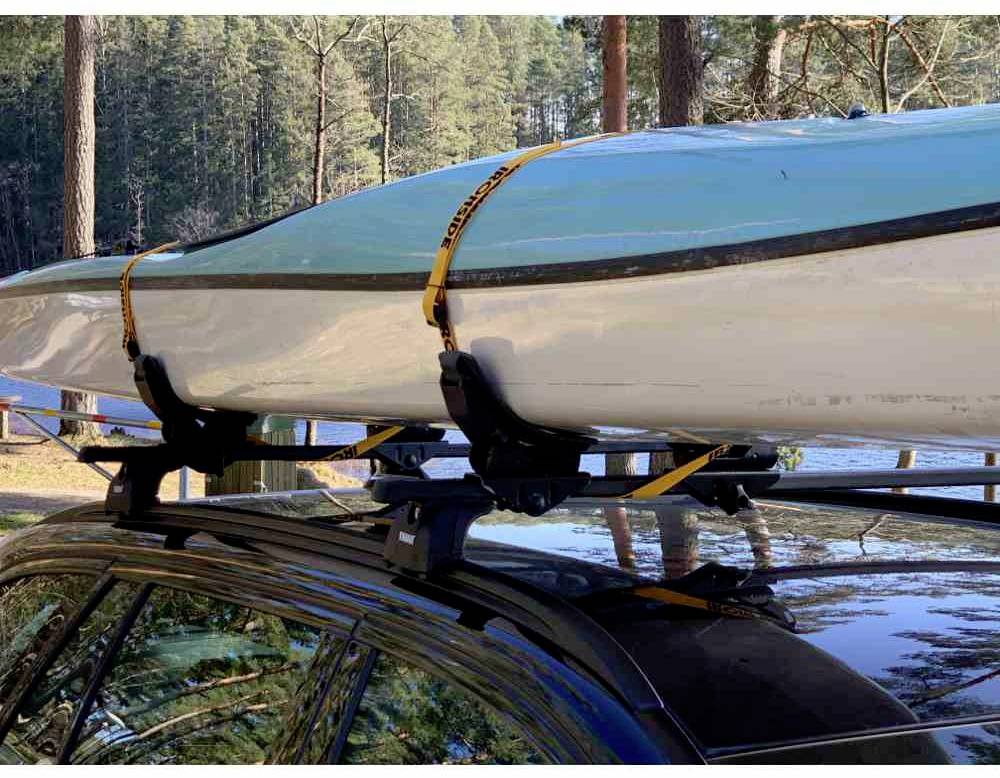 Kajak jest przymocowany pasami do dachu samochodu. W tle widać drzewa i jezioro, co wskazuje na otoczenie outdoorowe.