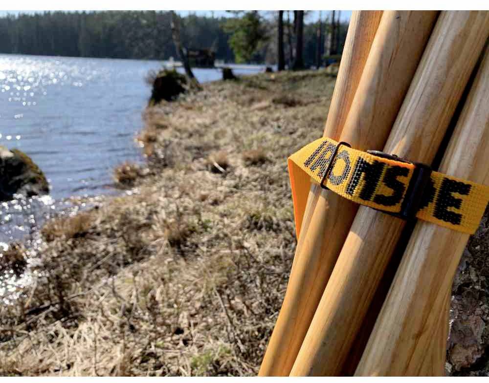 Drewniane kołki z pomarańczową wstążką na łące nad brzegiem jeziora. W tle widać wodę i drzewa.