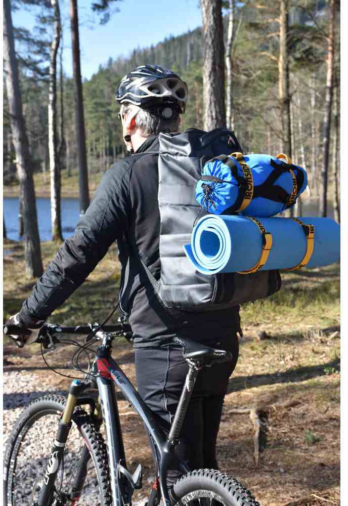 Osoba ubrana na czarno jedzie na rowerze w lesie. Na plecach ma duży plecak z niebieską matą izolacyjną i śpiworkiem.