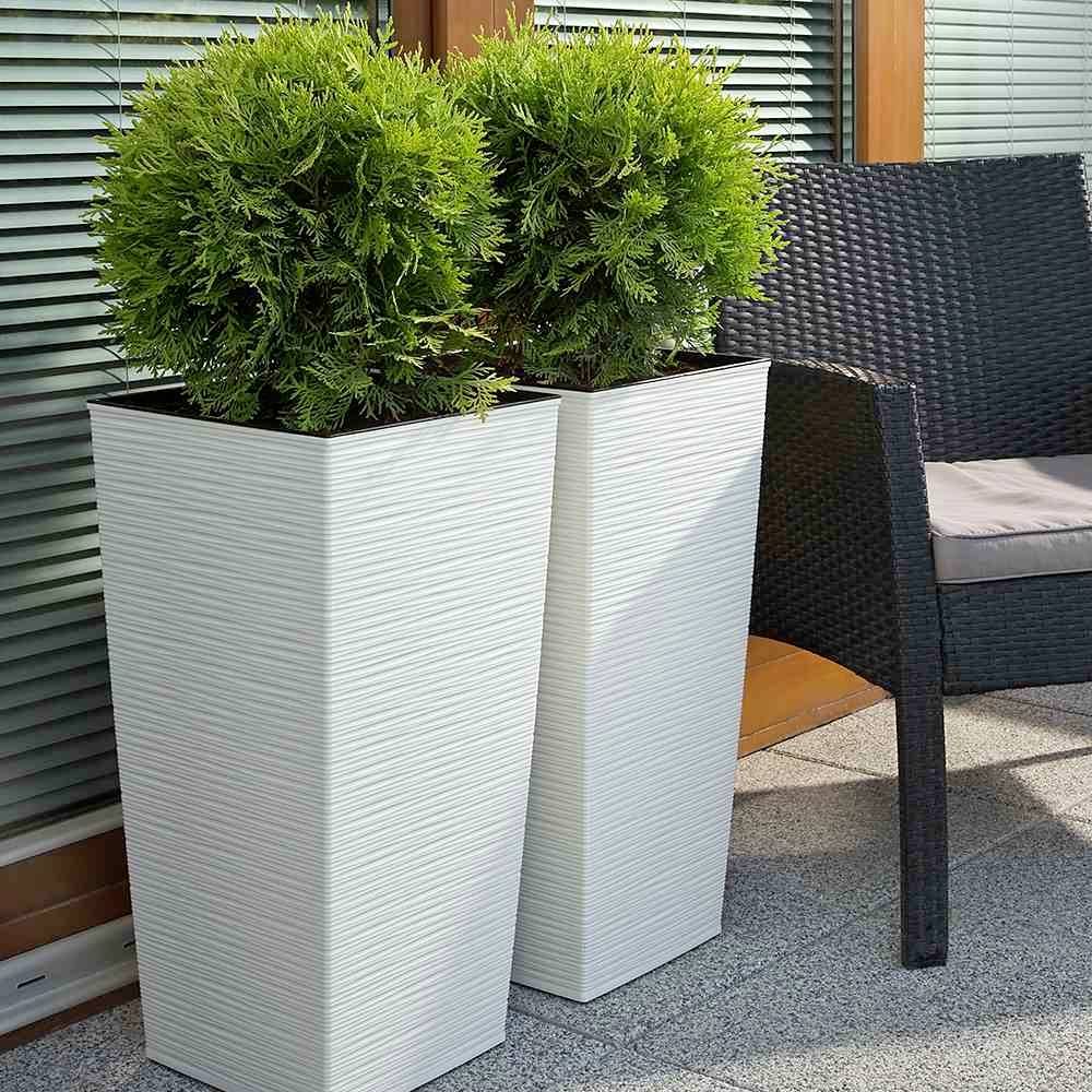 Dwa wysokie, prostokątne donice z jasnego materiału z zielonymi krzewami stoją na tarasie obok czarnego krzesła.