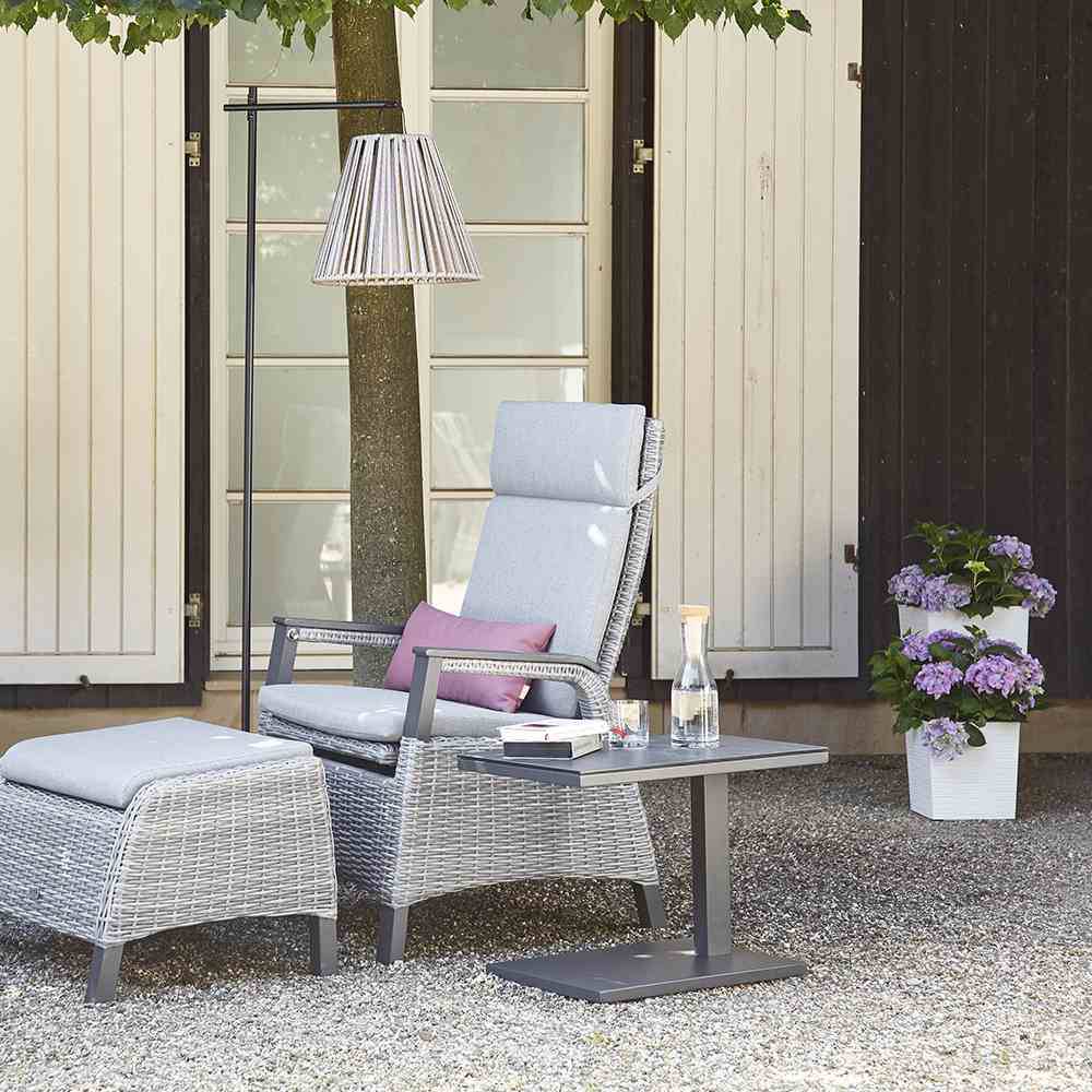 Fotel do użytku na świeżym powietrzu z podnóżkiem i stolikiem pomocniczym na żwirowej powierzchni przed drzewem. Obok stoi doniczka z fioletowymi kwiatami.