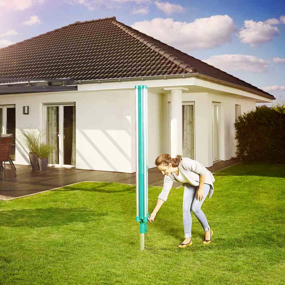 Kobieta instaluje niebieski parasol ogrodowy w soczystym zielonym ogrodzie przed nowoczesnym domem z ciemnym dachem i tarasem.