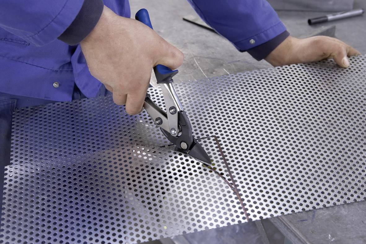 Osoba wycina kawałek blachy perforowanej za pomocą nożyc do cięcia metalu. Ręce trzymają nożyce i wykonują precyzyjne nacięcia w metalu.