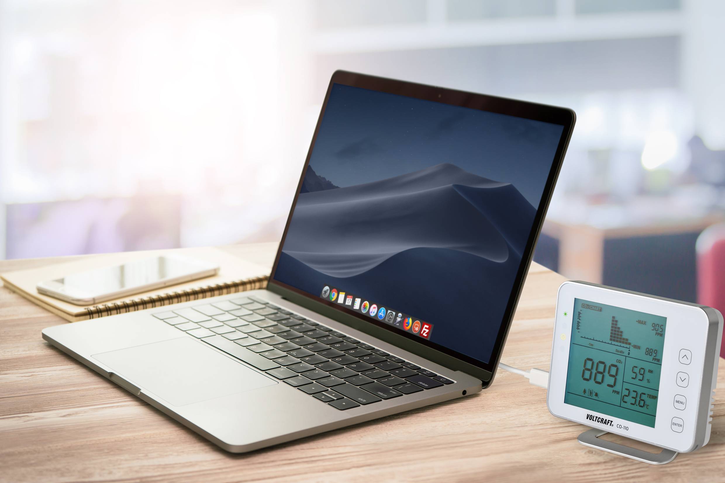 Laptop na biurku obok cyfrowego termo-higrometru i notesu. Na ekranie laptopa widoczny jest neutralny tło pulpitu.