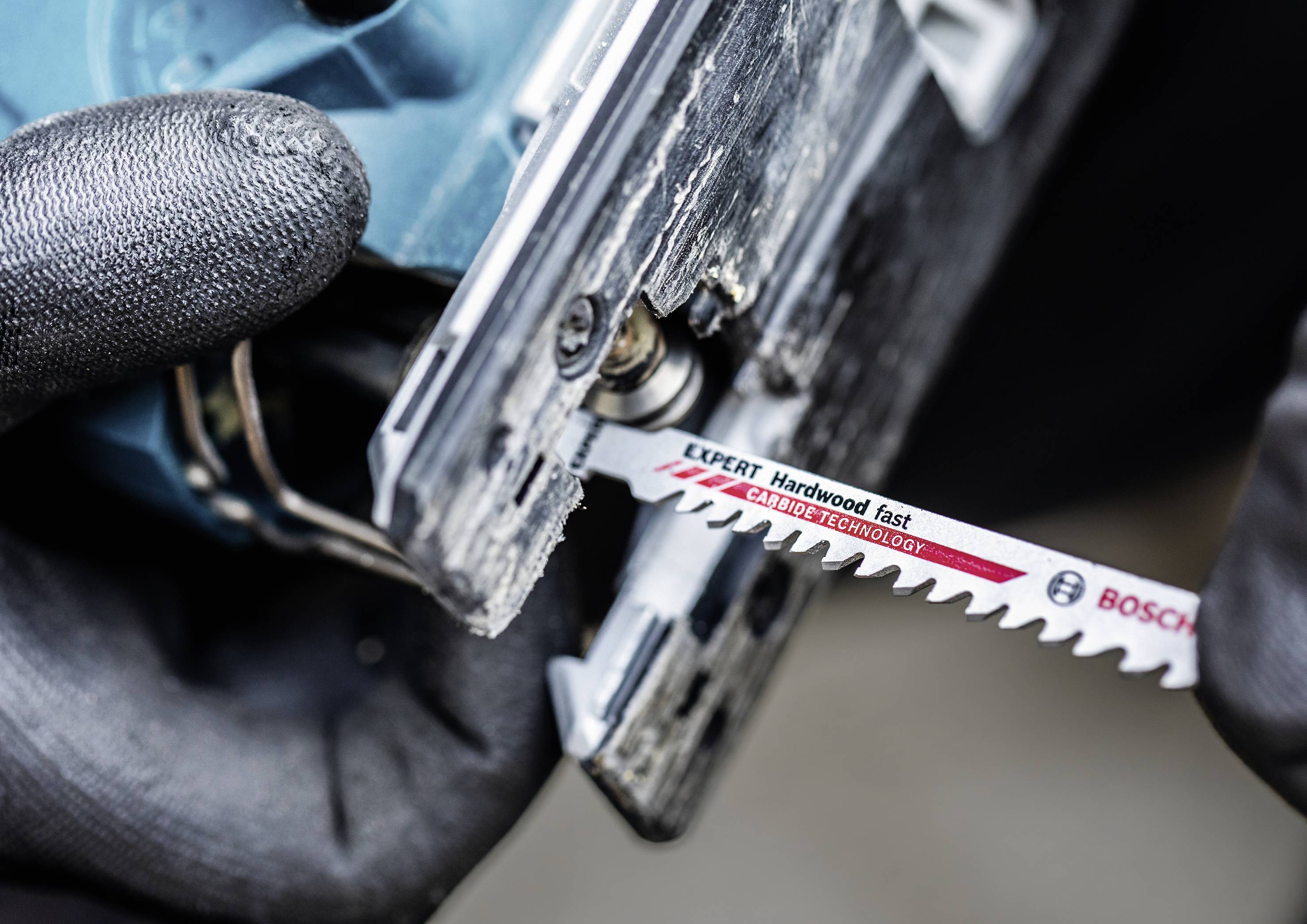 Czarna dłoń w rękawicy trzyma urządzenie elektryczne, podczas gdy druga wkłada brzeszczot Bosch z napisem 'Expert Hardwood fast'.