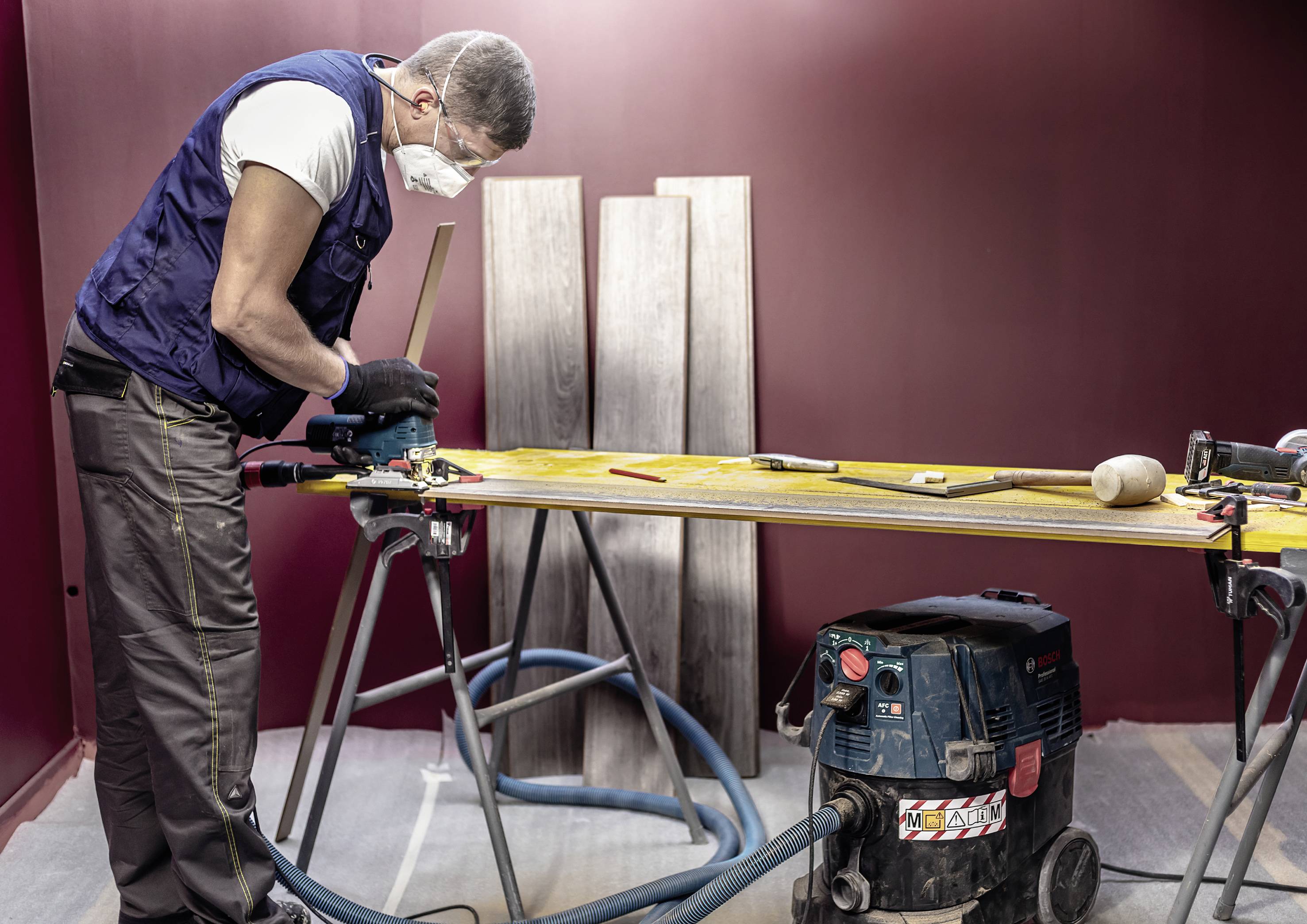 Mężczyzna w masce ochronnej pracuje w warsztacie stolarskim. Na stole leżą narzędzia i kawałki drewna. Obok niego stoi odkurzacz.