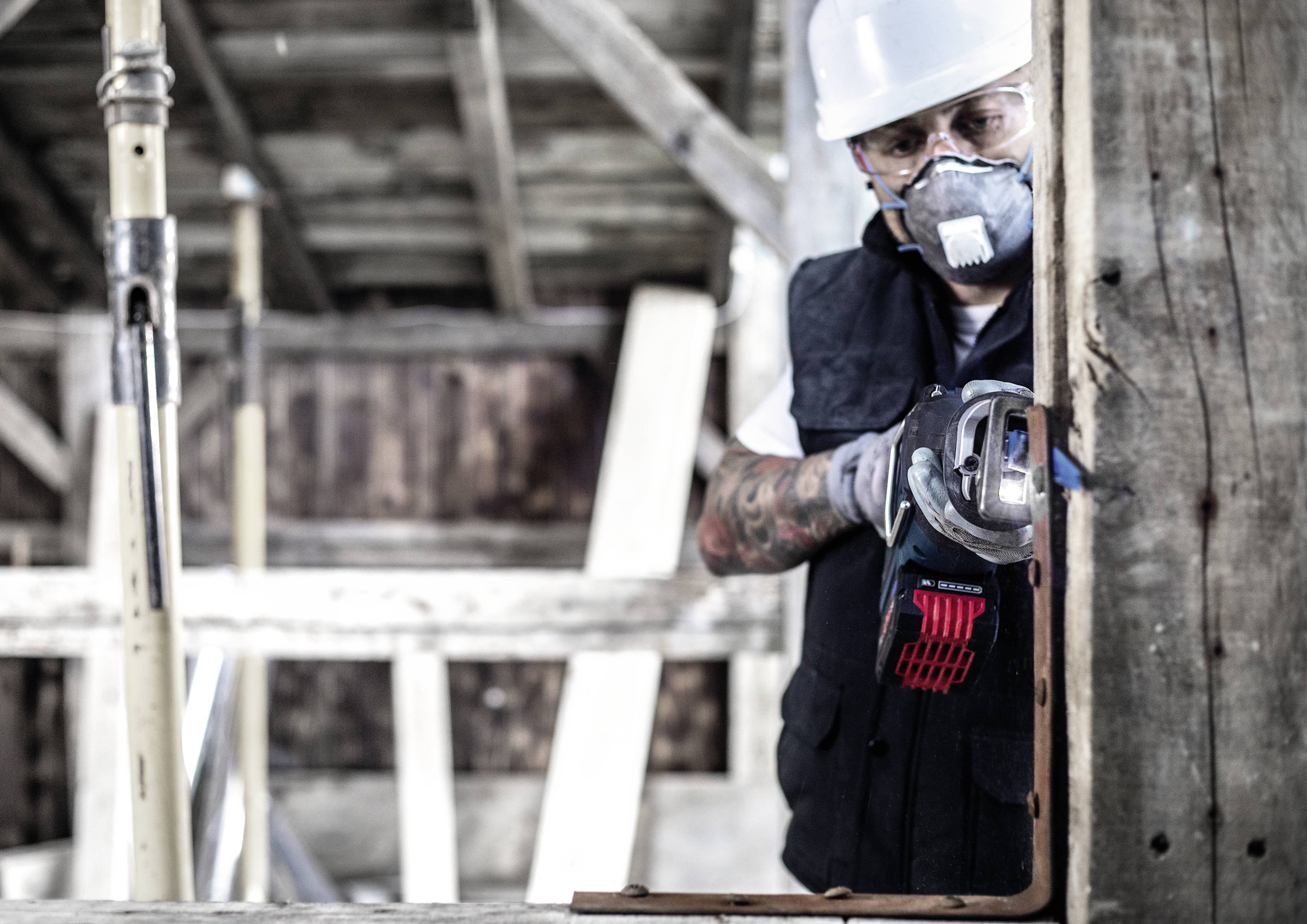 Robotnik budowlany w masce ochronnej i kasku używa elektronarzędzia do cięcia deski na terenie budowy.