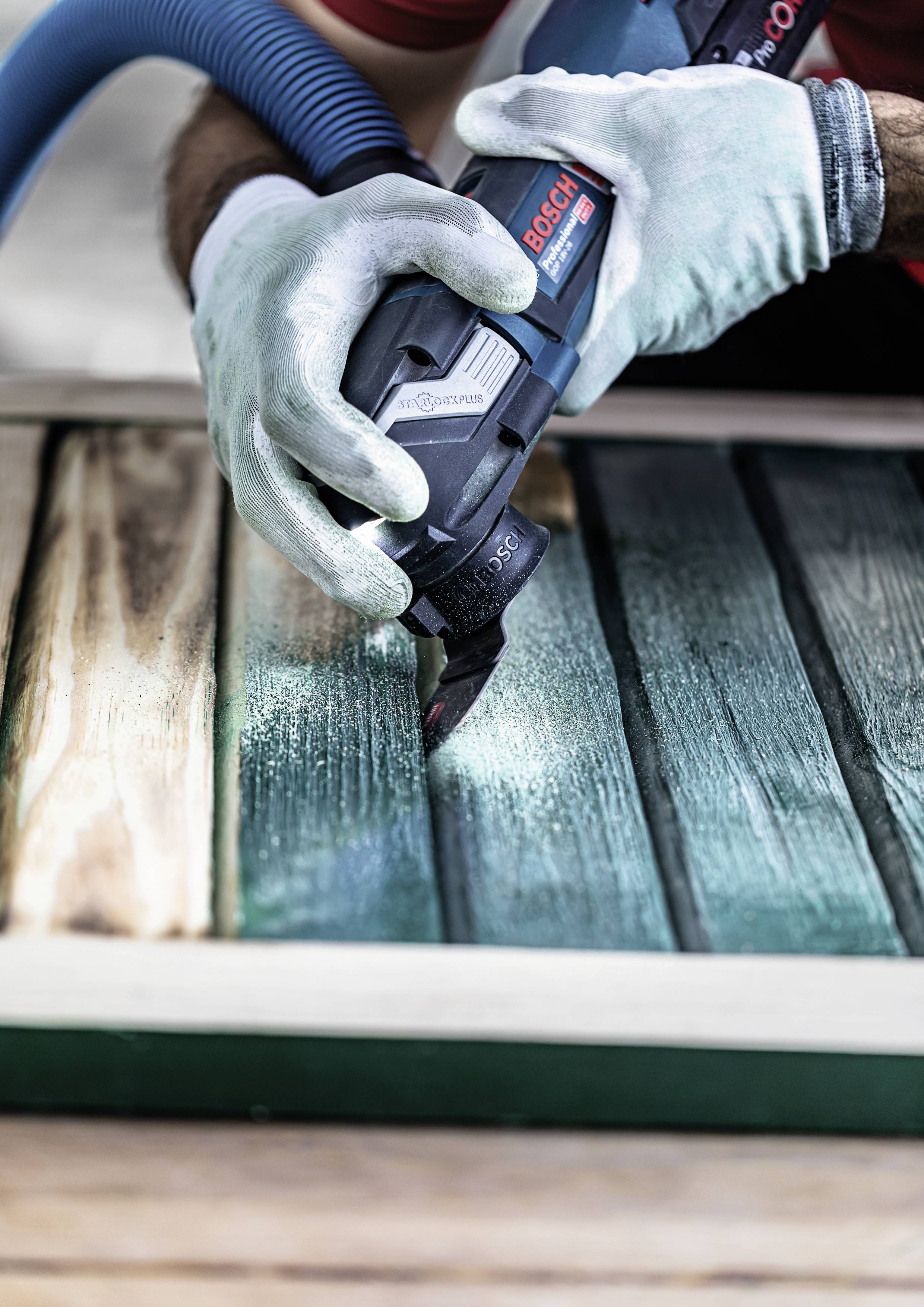 Mężczyzna szlifuje drewniane deski elektronarzędziem. Nosi niebieskie okulary ochronne i rękawice; obrabia drewno o zielonkawym odcieniu.