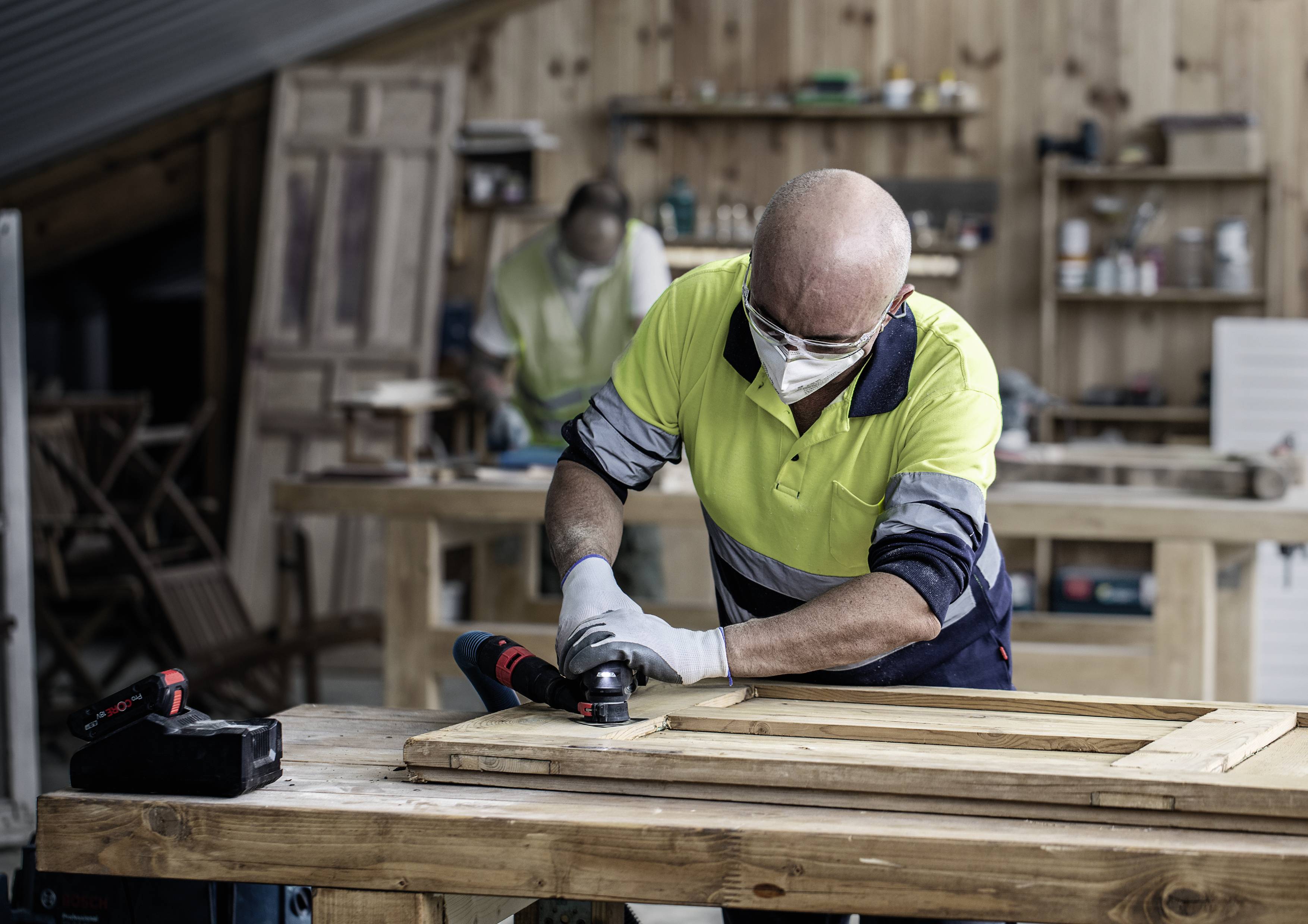 Mężczyzna w odzieży ochronnej pracuje elektronarzędziem przy desce drewnianej w warsztacie. W tle widać kolejną osobę.