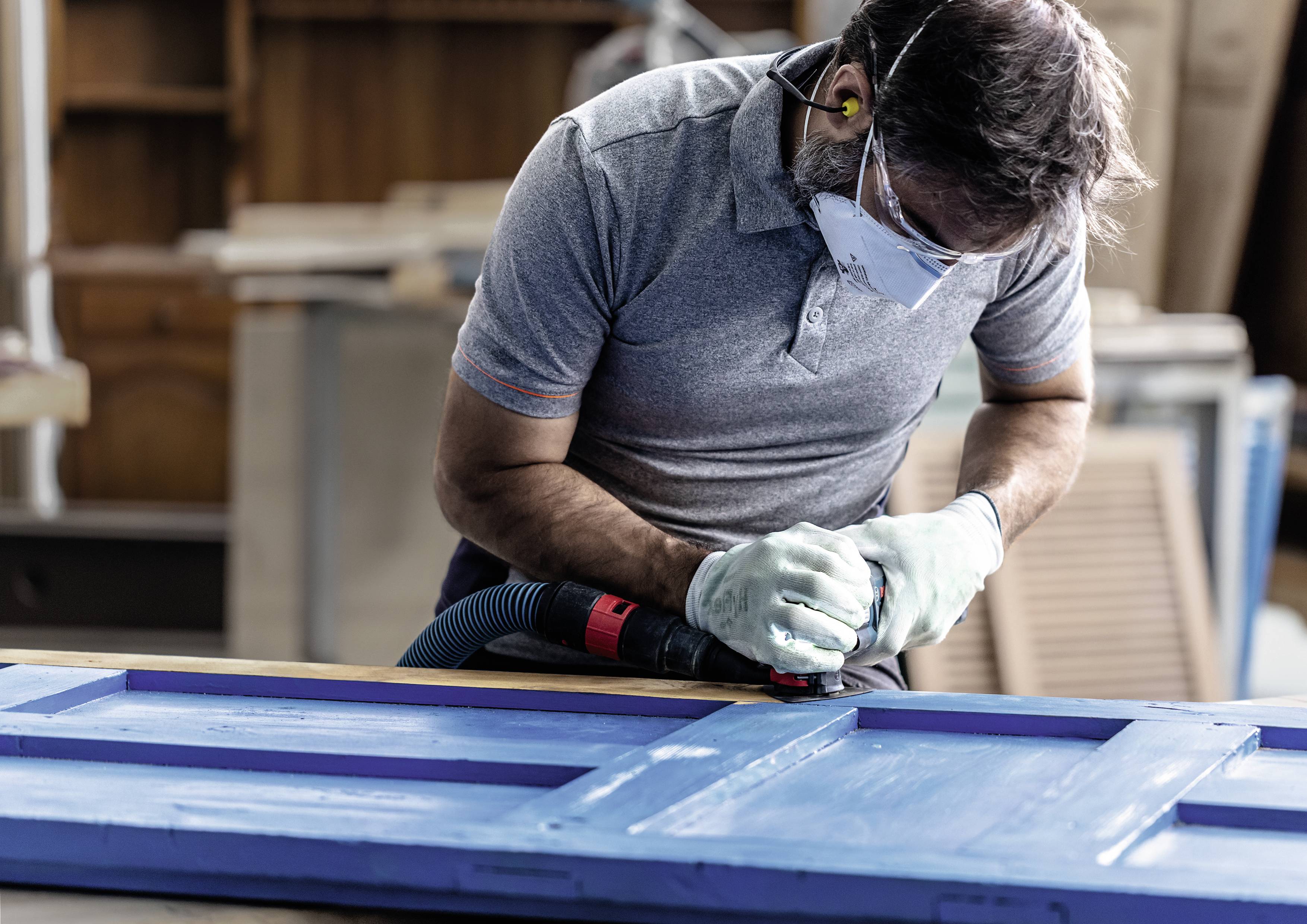 Mężczyzna szlifuje niebieską powierzchnię drewna za pomocą szlifierki. Nosi rękawice ochronne, okulary i maskę przeciwpyłową w warsztacie.