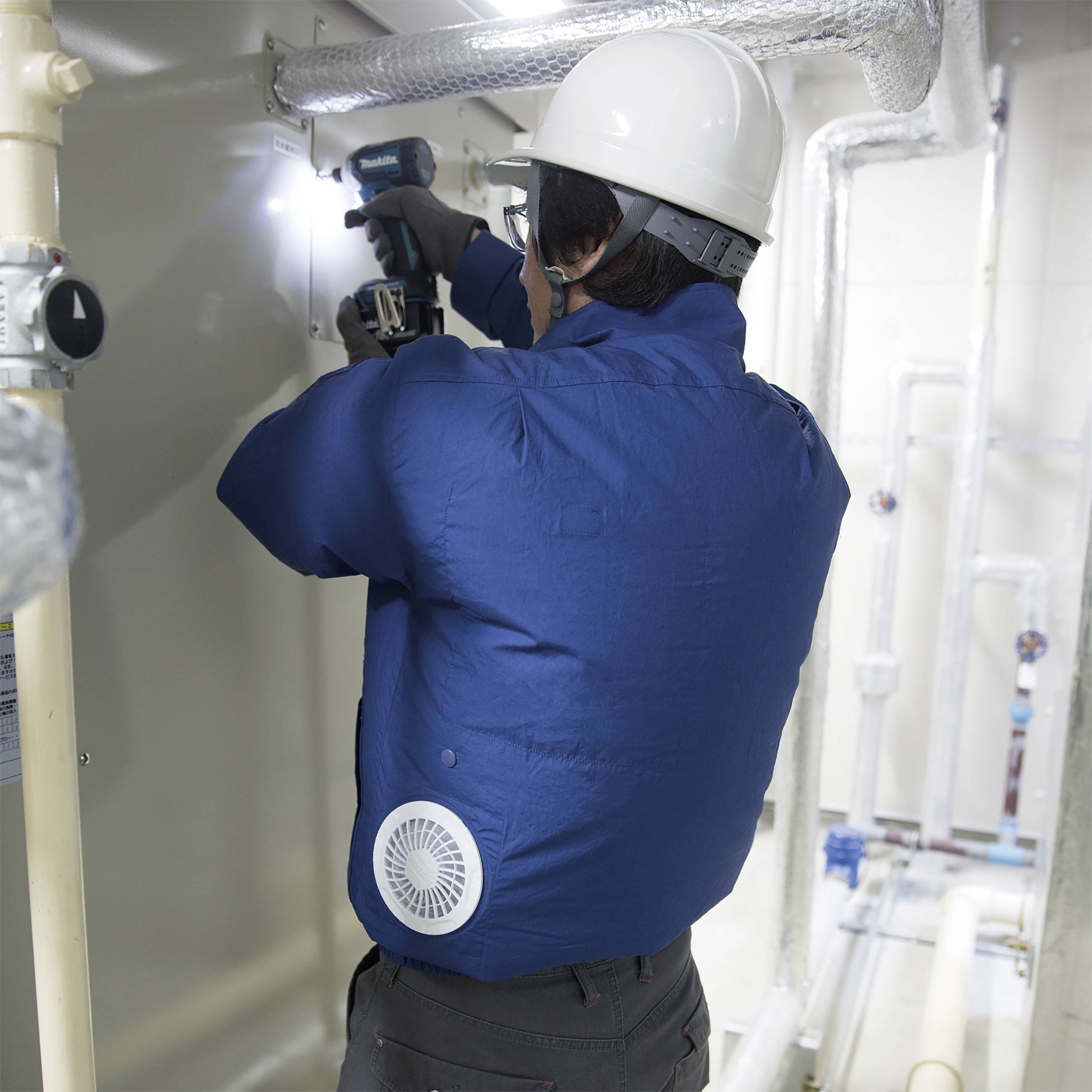 Robotnik w kasku ochronnym i niebieskiej kurtce mocuje rury wkrętarką akumulatorową w pomieszczeniu technicznym.