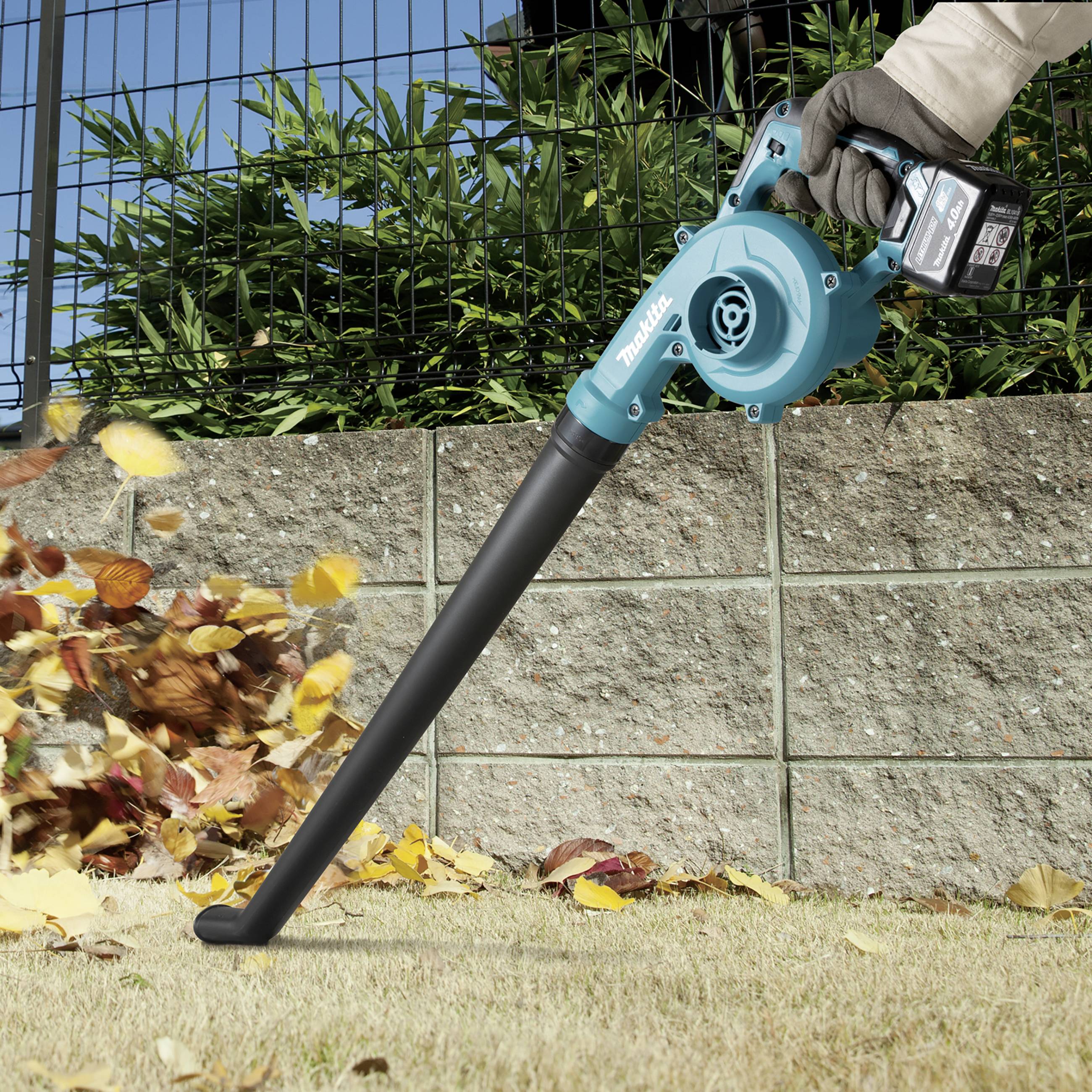 Osoba używa zasilanego baterią dmuchawy do liści, aby zdmuchiwać liście w jednym kierunku na trawniku. Na tylnym planie widoczny jest płot.