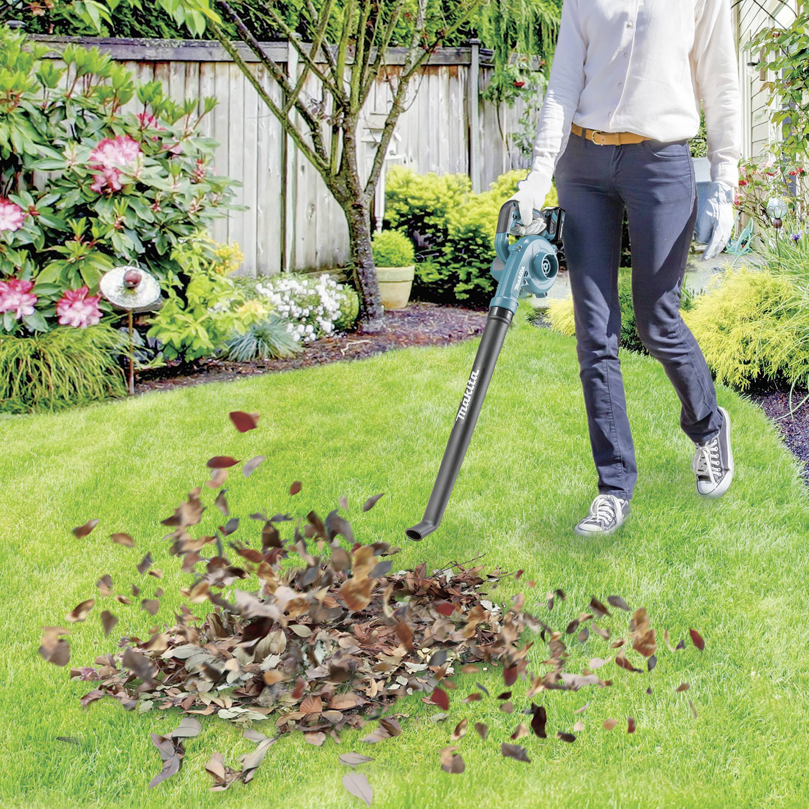 Osoba stoi w ogrodzie i używa dmuchawy do liści, aby zebrać liście na trawniku. W tle widoczne są rośliny i drewniany płot.