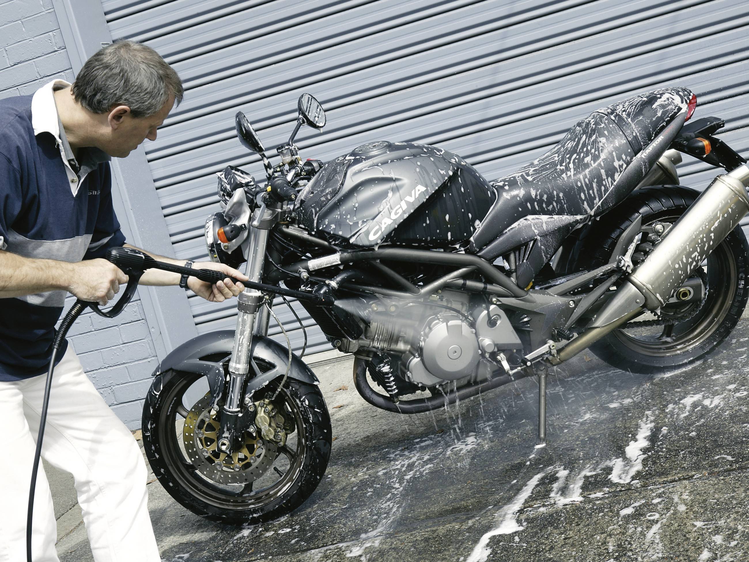 Mężczyzna myje motocykl myjką ciśnieniową, pokryty pianą mydlaną. W tle zamknięta roleta.
