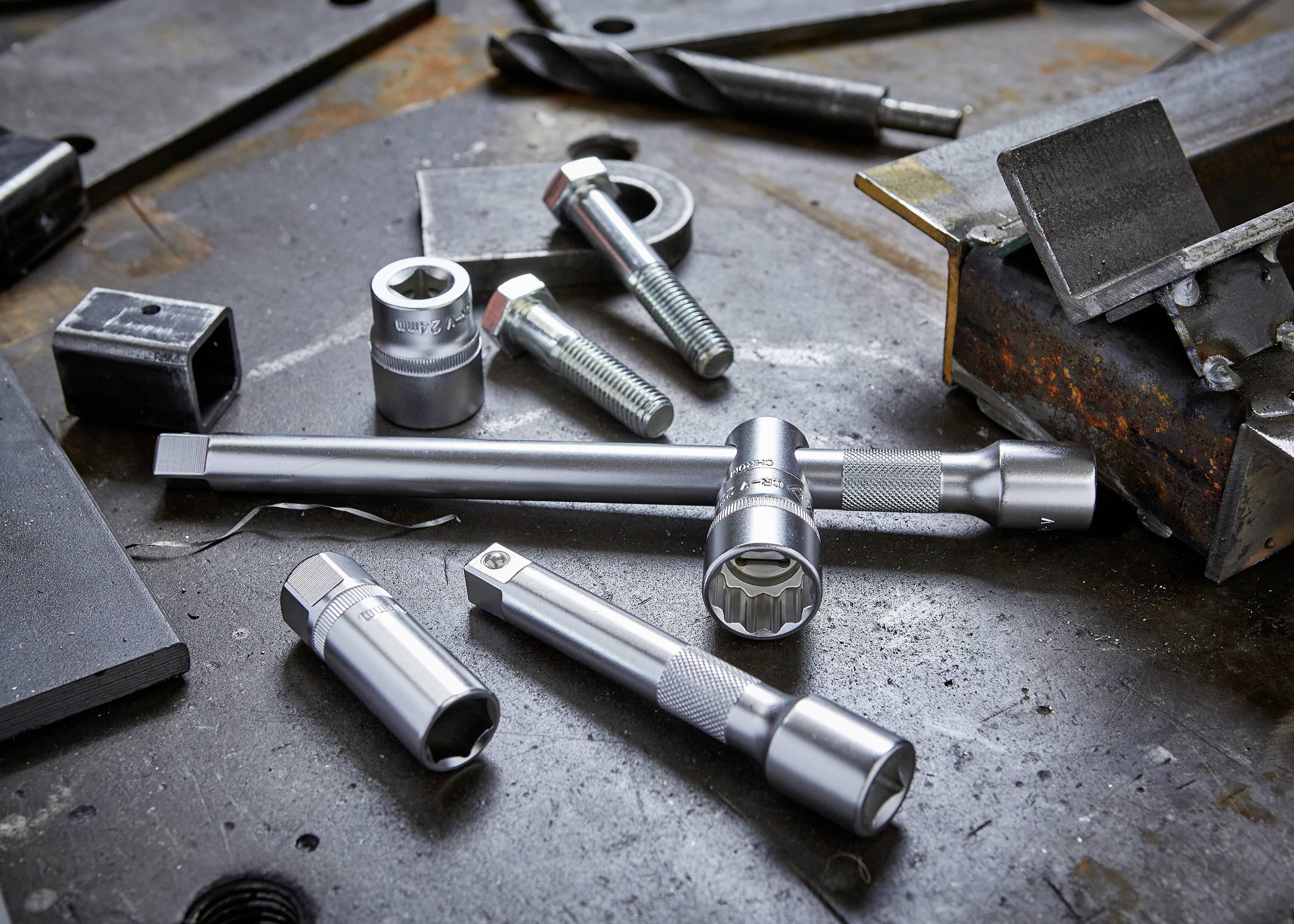 Na metalowym stole leżą różne narzędzia, takie jak klucze nasadowe i śruby, w tle widoczne są stalowe płyty i wiertła.