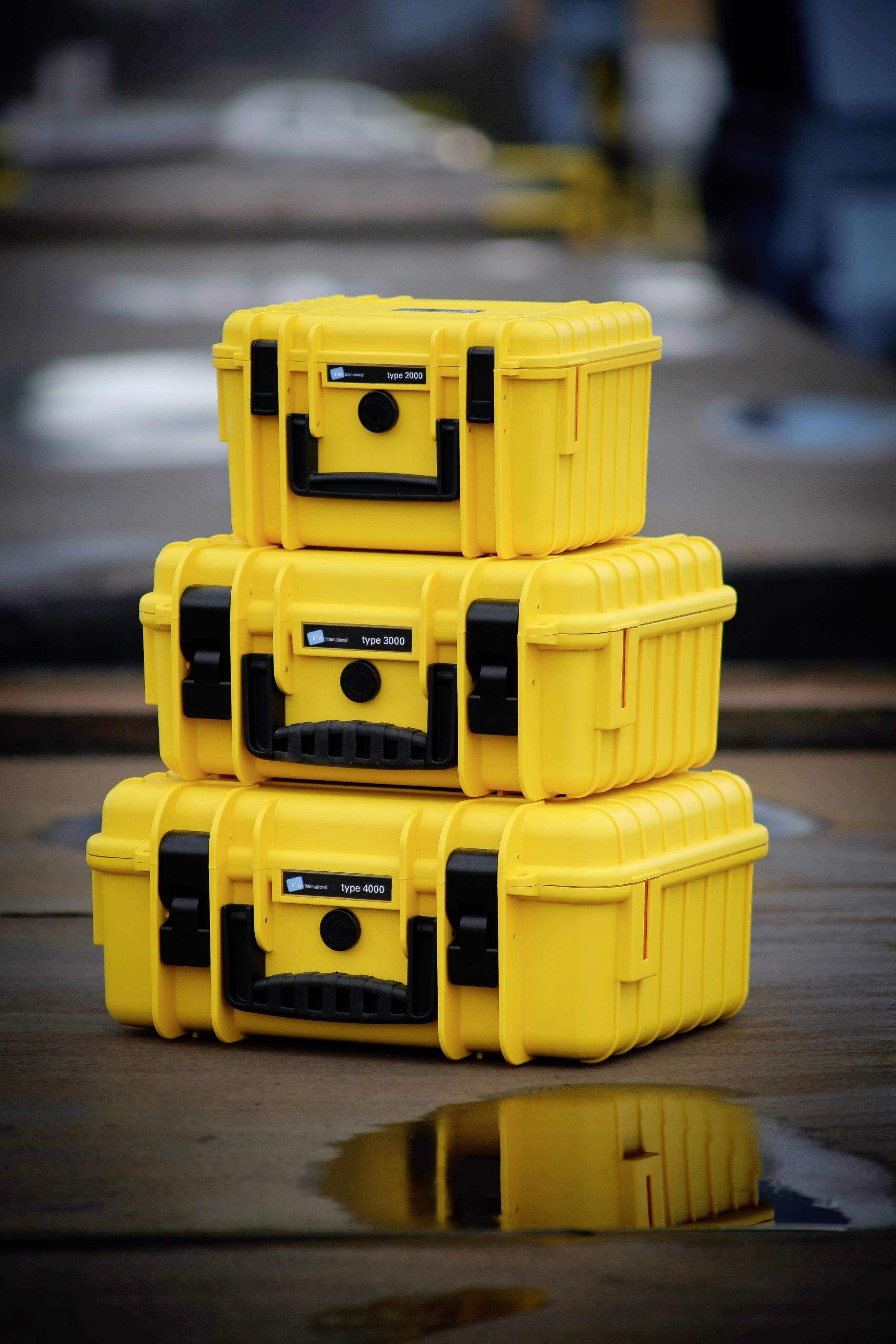Trzy żółte, wytrzymałe walizki z tworzywa sztucznego są ułożone jedna na drugiej. Stoją na drewnianym podłożu. Tło jest utrzymane w stylu przemysłowym.