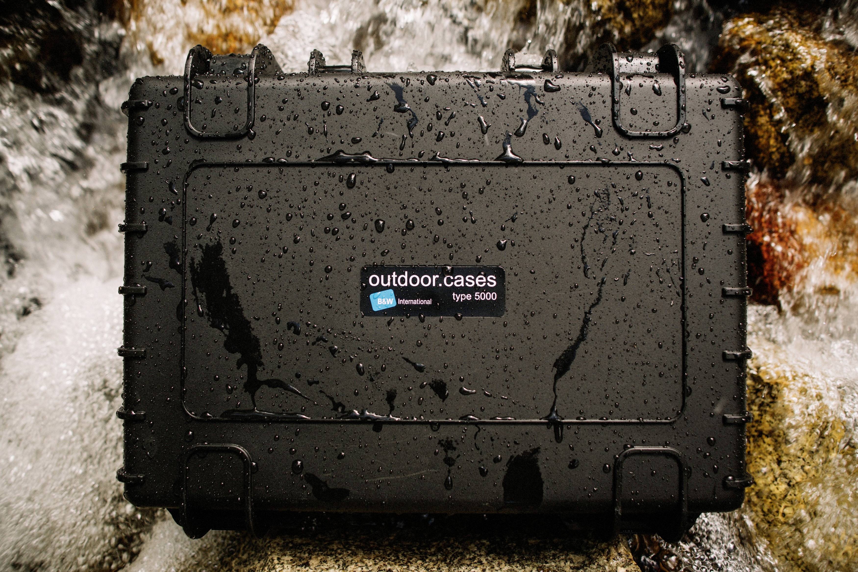 Wodoodporny czarny futerał ochronny z napisem 'outdoor.cases type 5000' leży na mokrych kamieniach w wodzie.