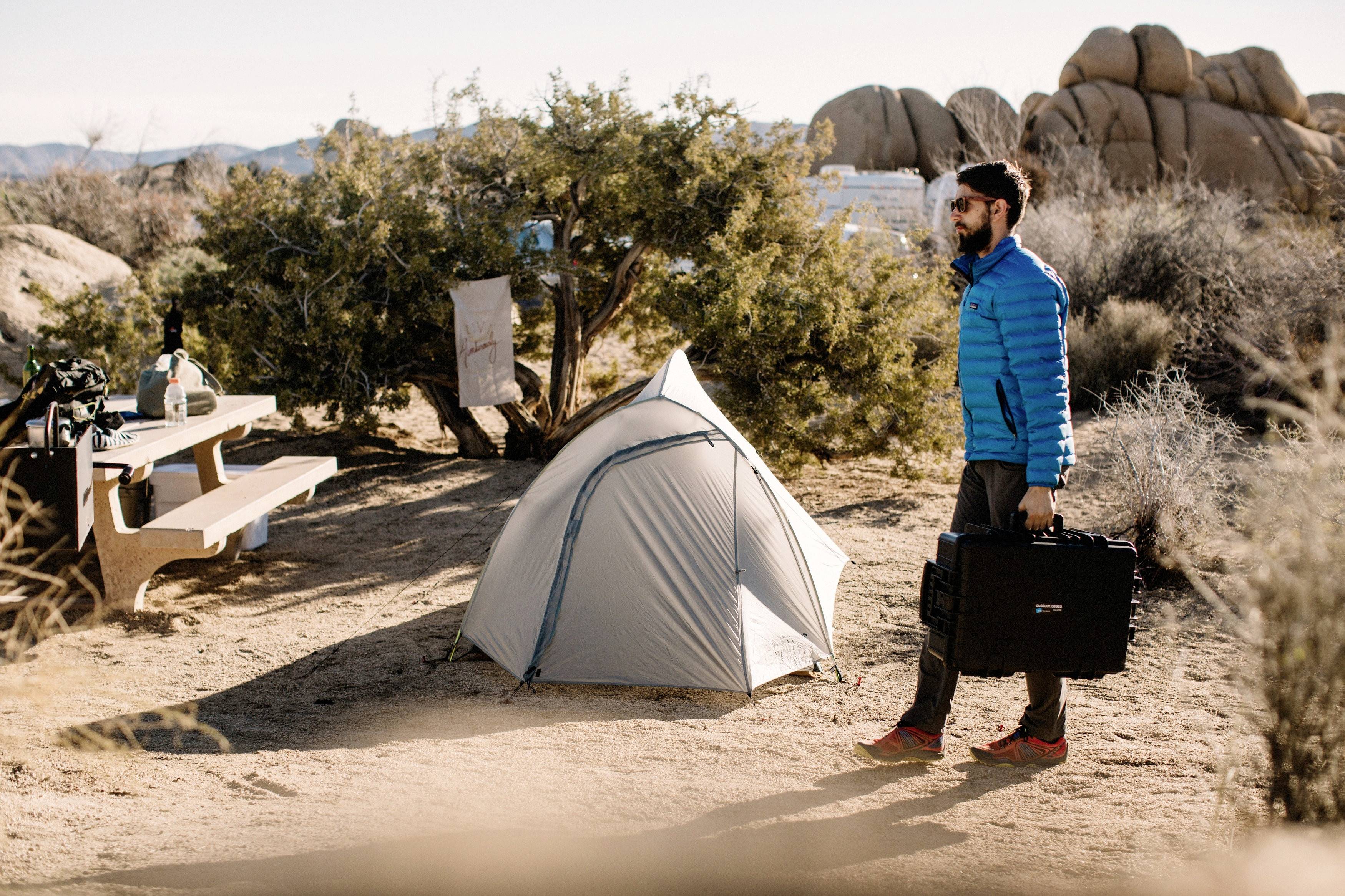 Mężczyzna w niebieskiej kurtce niesie walizkę na kempingu, pośród namiotów i drzew; słoneczny dzień z skałami w tle.