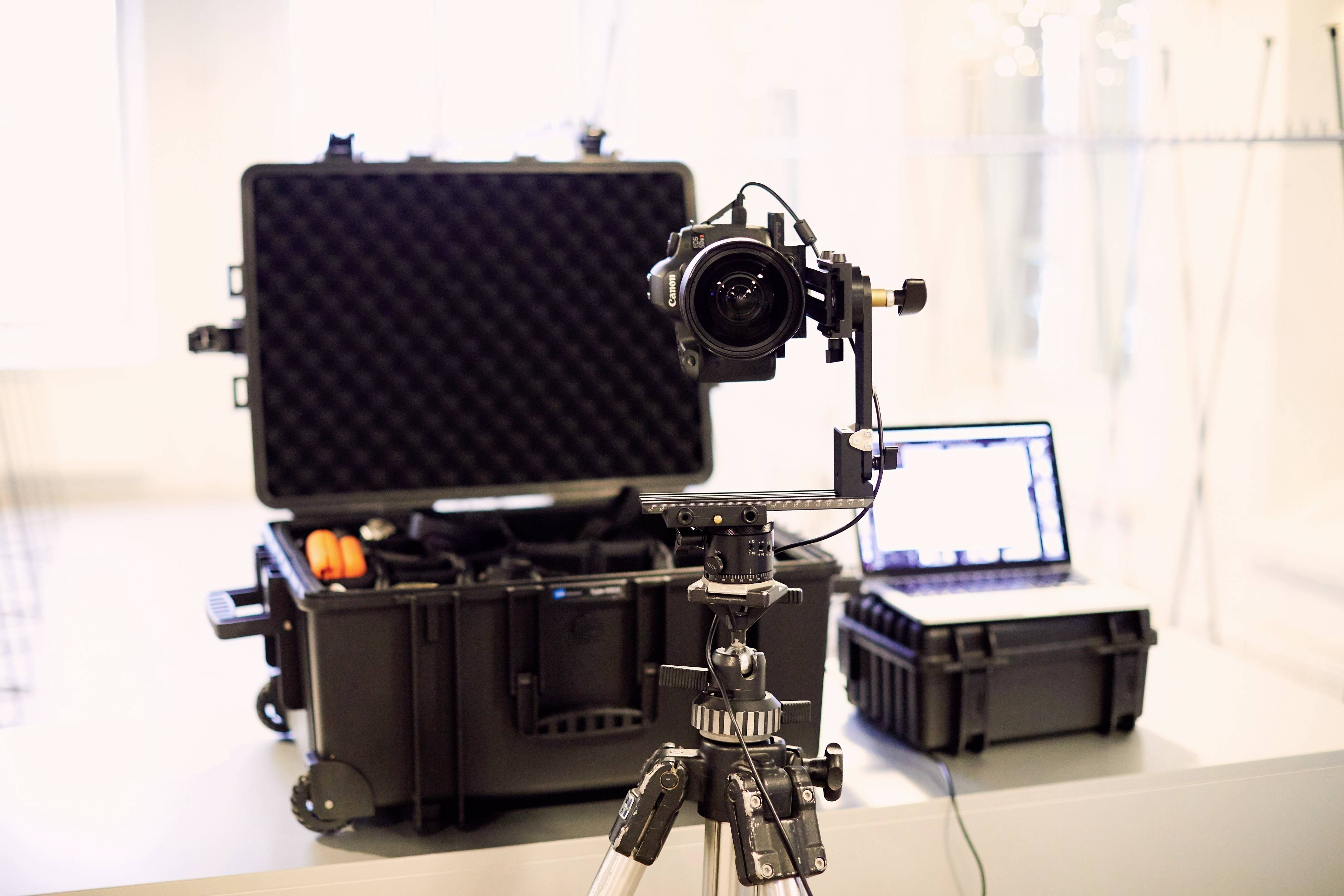 Kamera na statywie przed otwartą walizką i laptopem; przygotowanie do zdjęcia lub nagrania wideo.