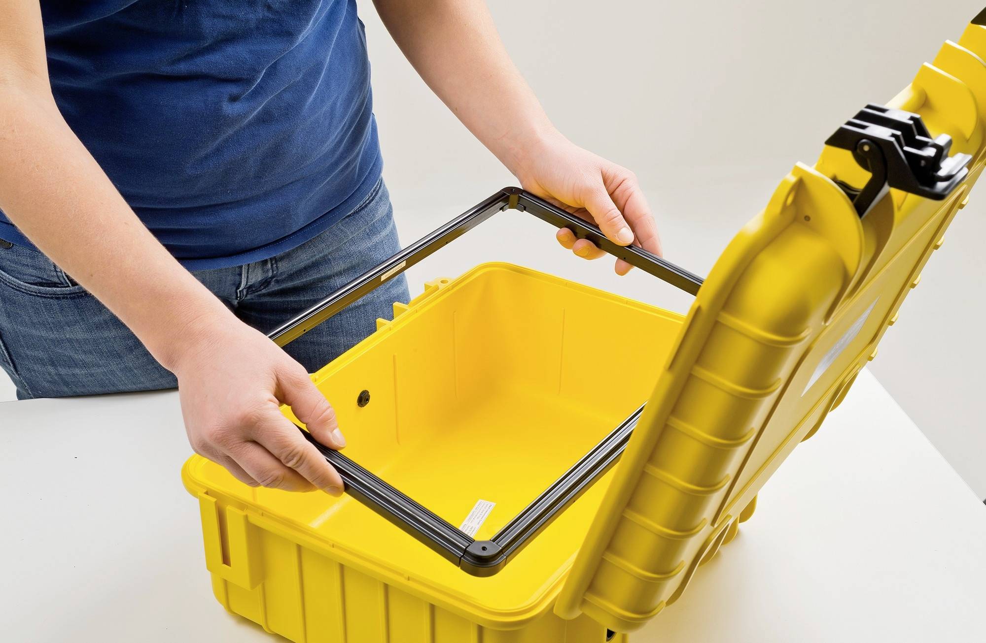 Osoba otwiera żółty, wytrzymały kufer na narzędzia wykonany z tworzywa sztucznego. Kufer posiada czarną uszczelkę w pokrywie.