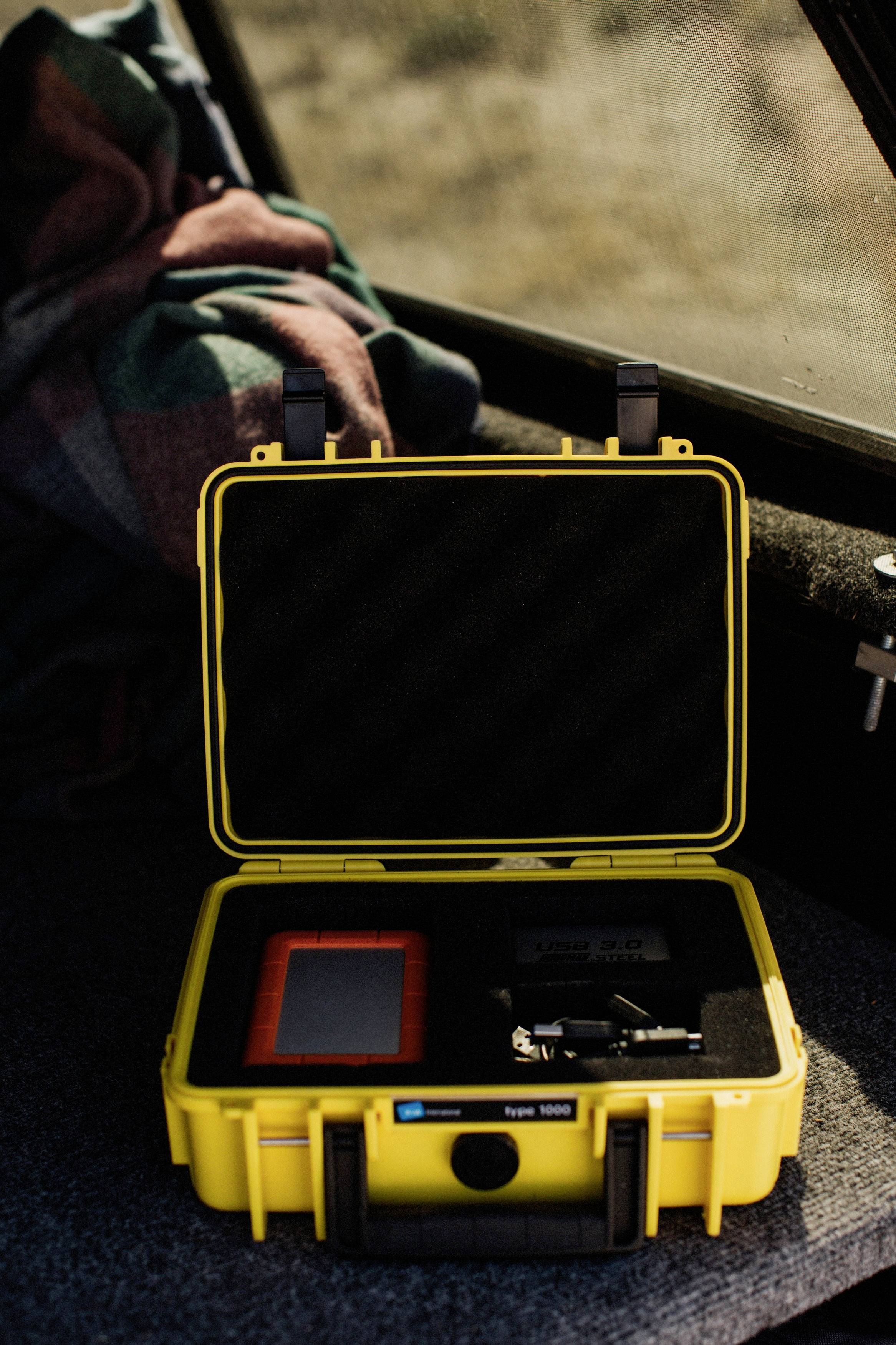 Żółta twarda walizka z otwartą pokrywą, w której znajduje się urządzenie w kolorach czerwonym i czarnym oraz dwie małe czarne części dodatkowe, umieszczona w tylnej części samochodu.