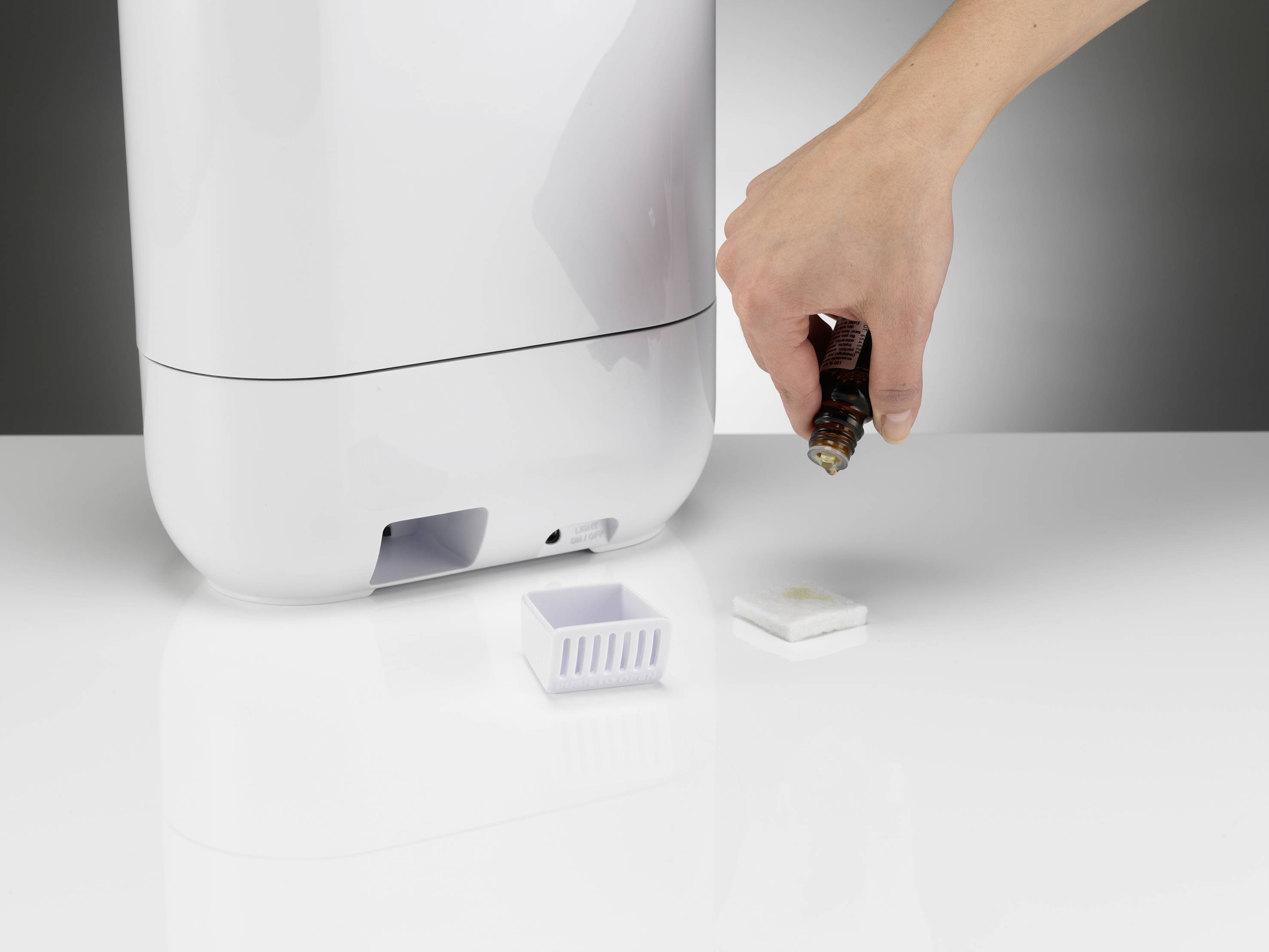 Dłoń zakrapla olejek eteryczny na wkład obok nawilżacza powietrza. Małe przegródka jest otwarta, aby przyjąć wkład.