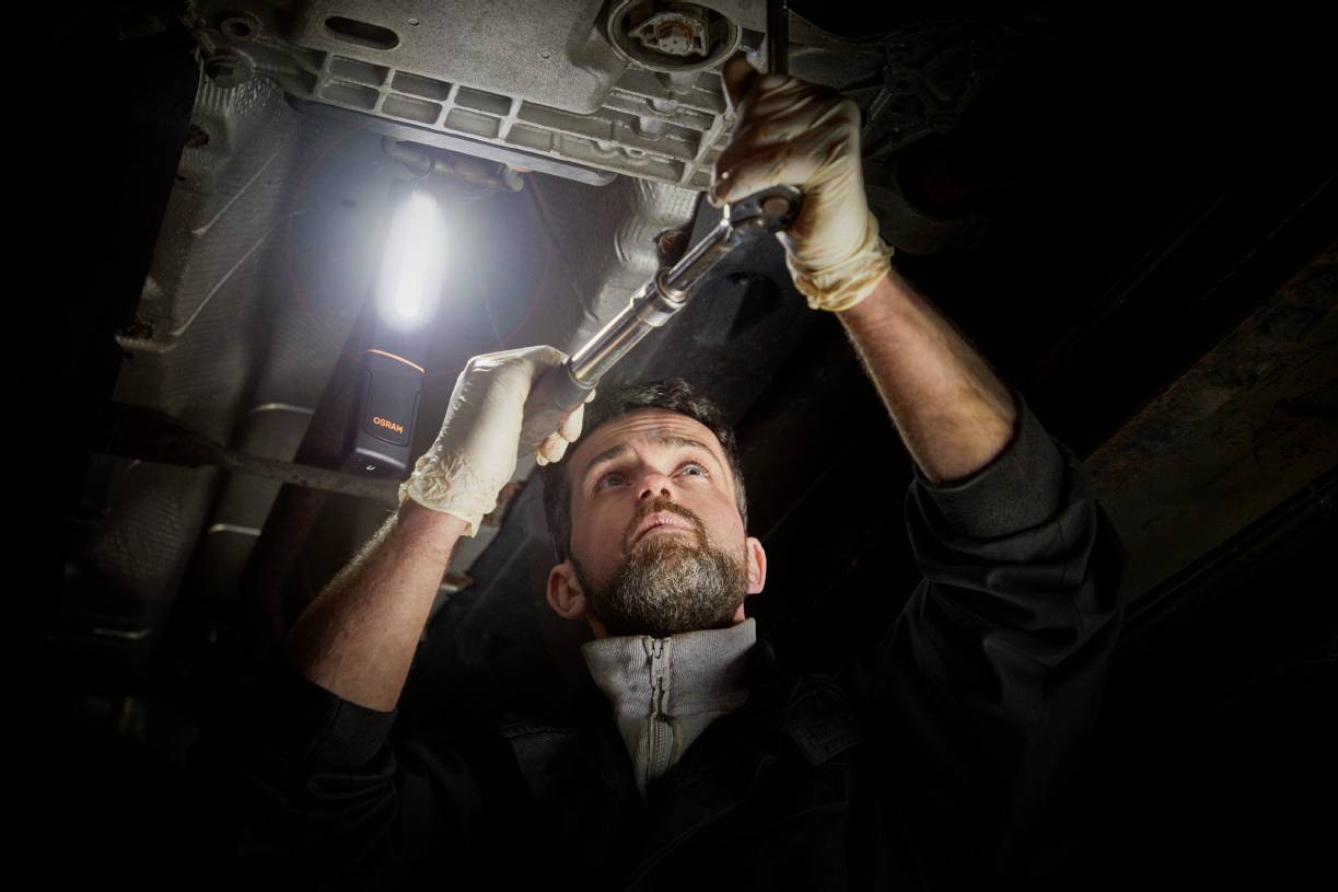 Mechanik pracuje pod samochodem, trzyma klucz płaski i jest ubrany w rękawice. Lampa oświetla jego obszar pracy.